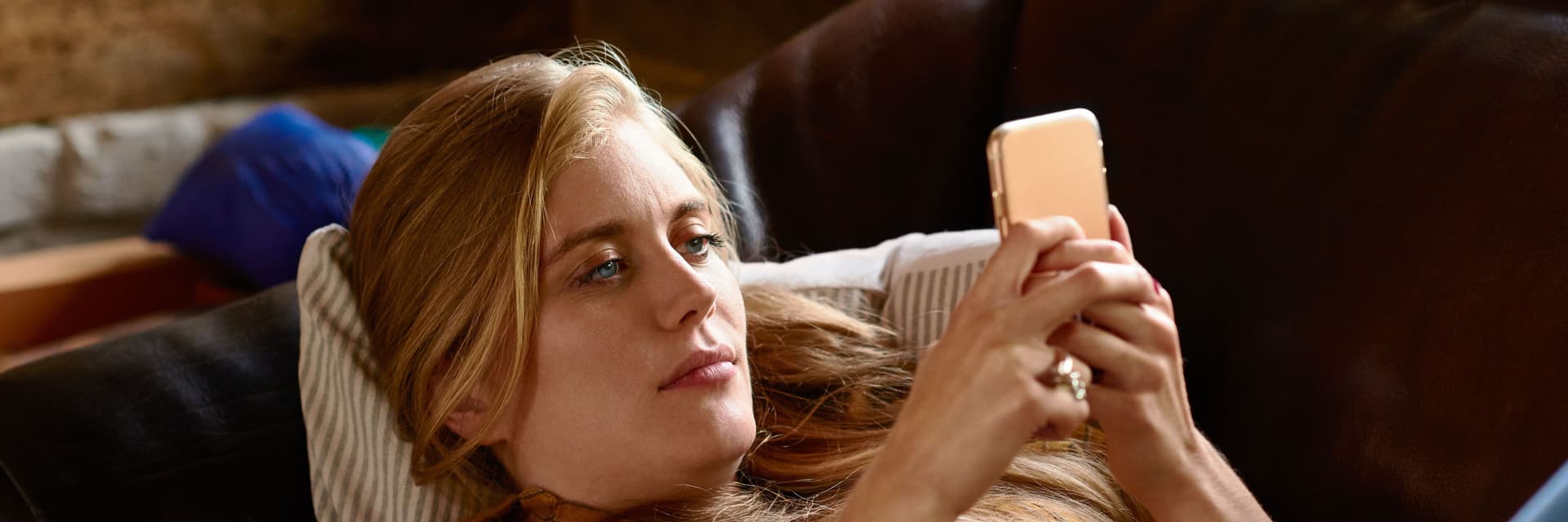 Eine blonde Frau liegt mit ihrem Handy in der Hand auf einem Sofa.