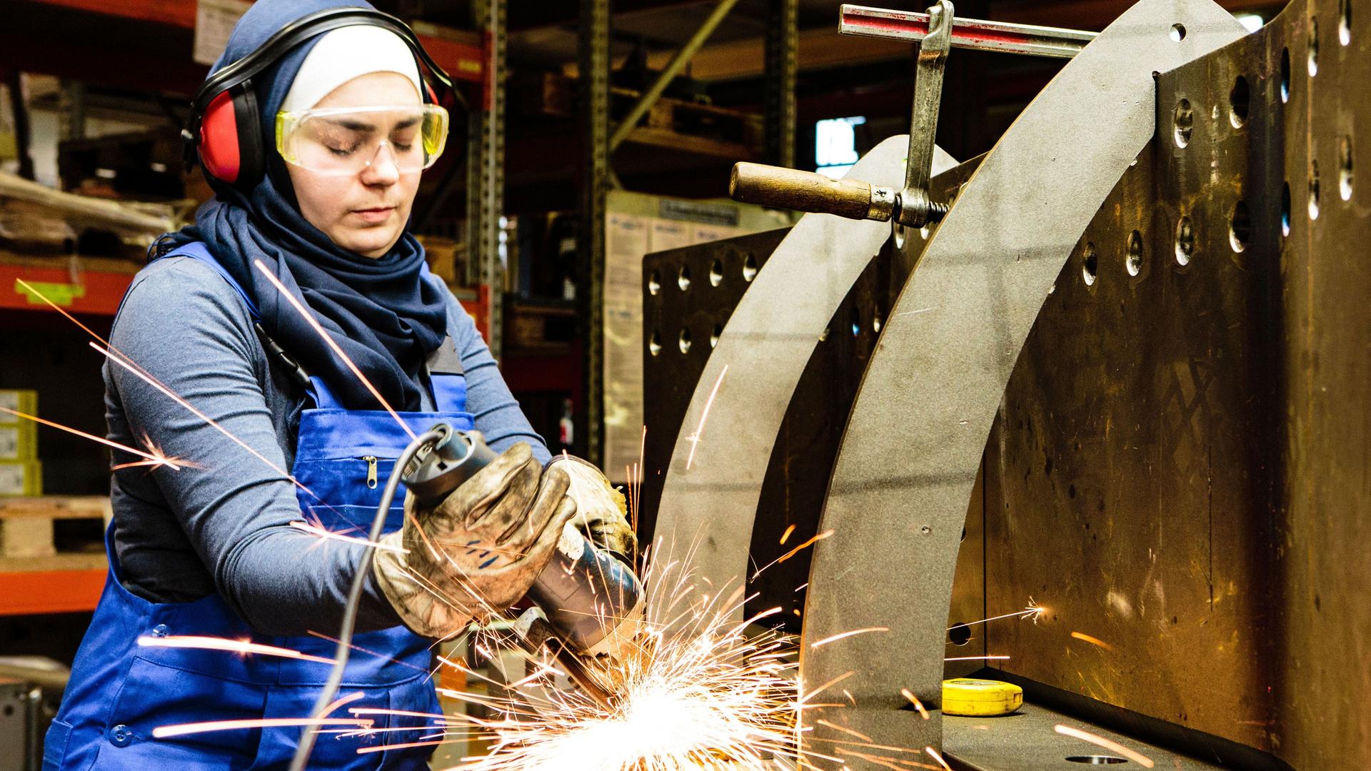 Eine junge Frau mit Kopftuch steht in einer Metall-Werkstatt. Sie arbeitet an einem Werkstück.