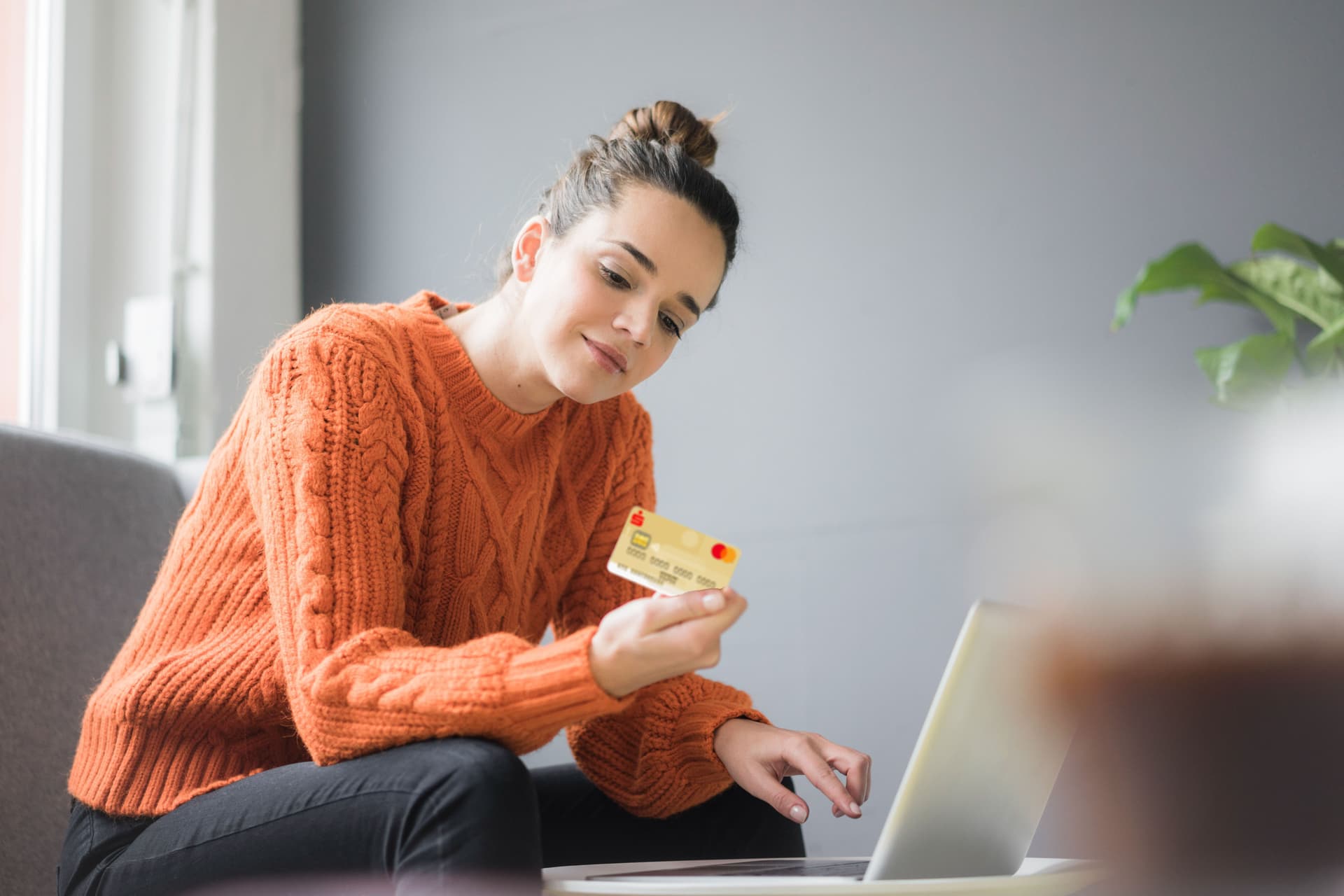 Eine junge Frau in orangenen Pulli sitzt vor einem Laptop. In der Hand hat sie eine goldene Kreditkarte. Sie schaut auf die Rückseite der Kreditkarte.