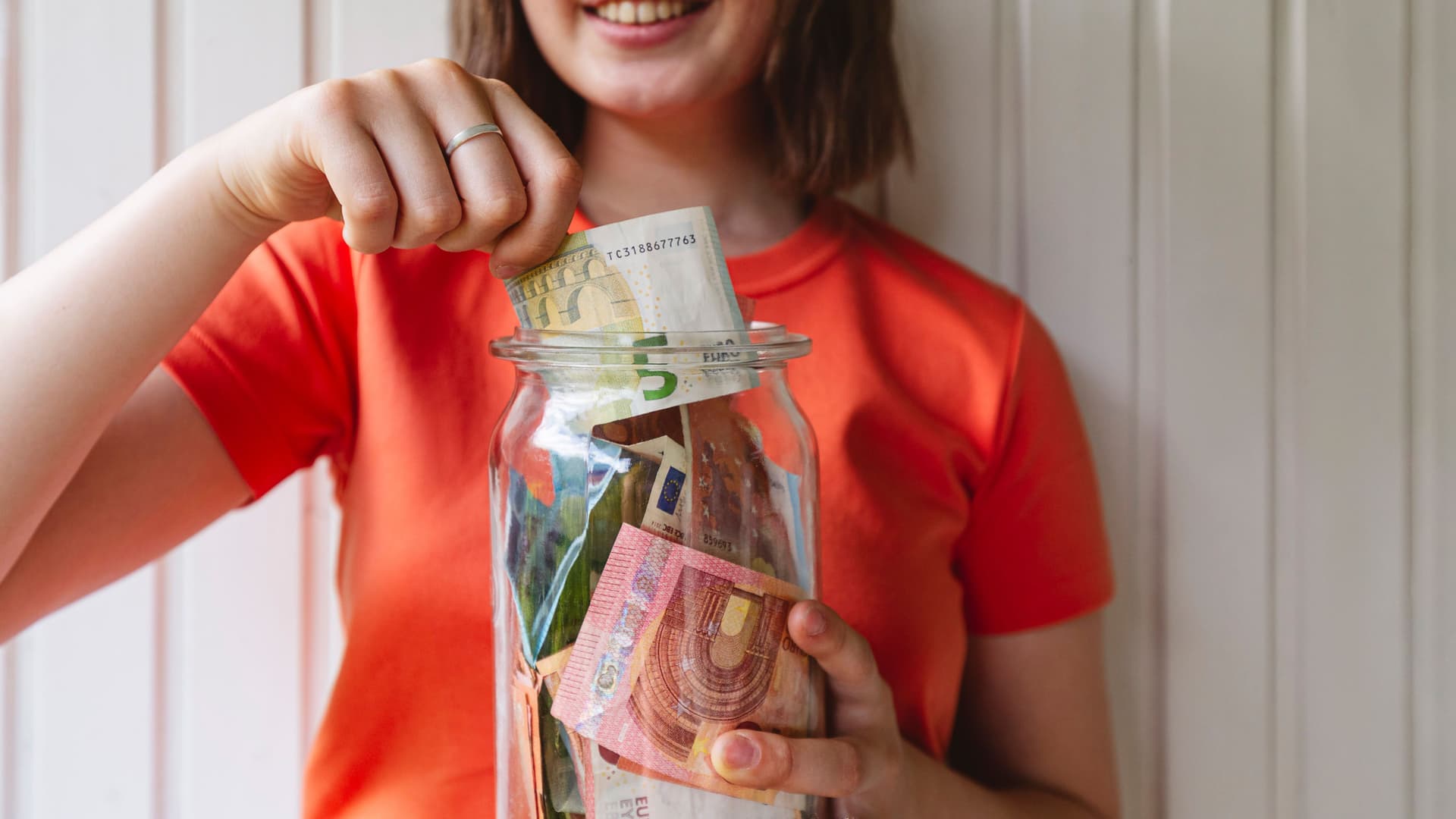 Oberkörper einer jungen Frau mit einem Glas voller Geld vor sich. mit einer Hand steckt sie Geld ins Glas.