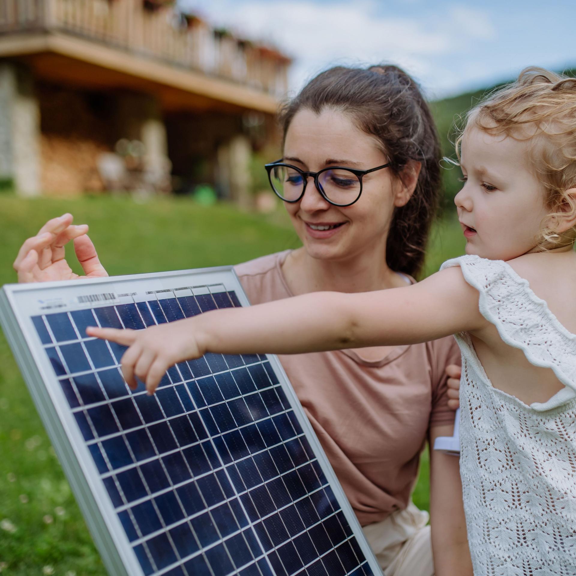 Eine junge Frau zeigt ihrer Tochter ein Solarpanel. Sie stehen auf einer Wiese. Im Hintergrund ist ein Haus zu sehen.