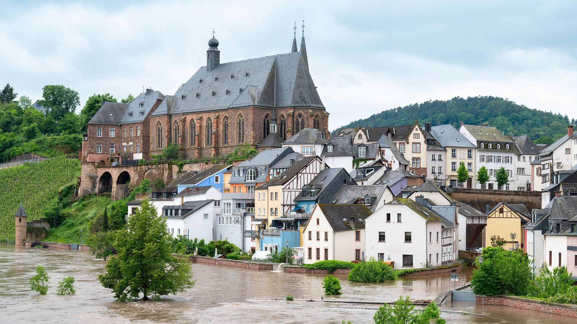 Ein überflutete Stadt am Fluss. Auf einem Hügel steht eine Kirche. Davor befinden sich überflutete Wohnhäuser.