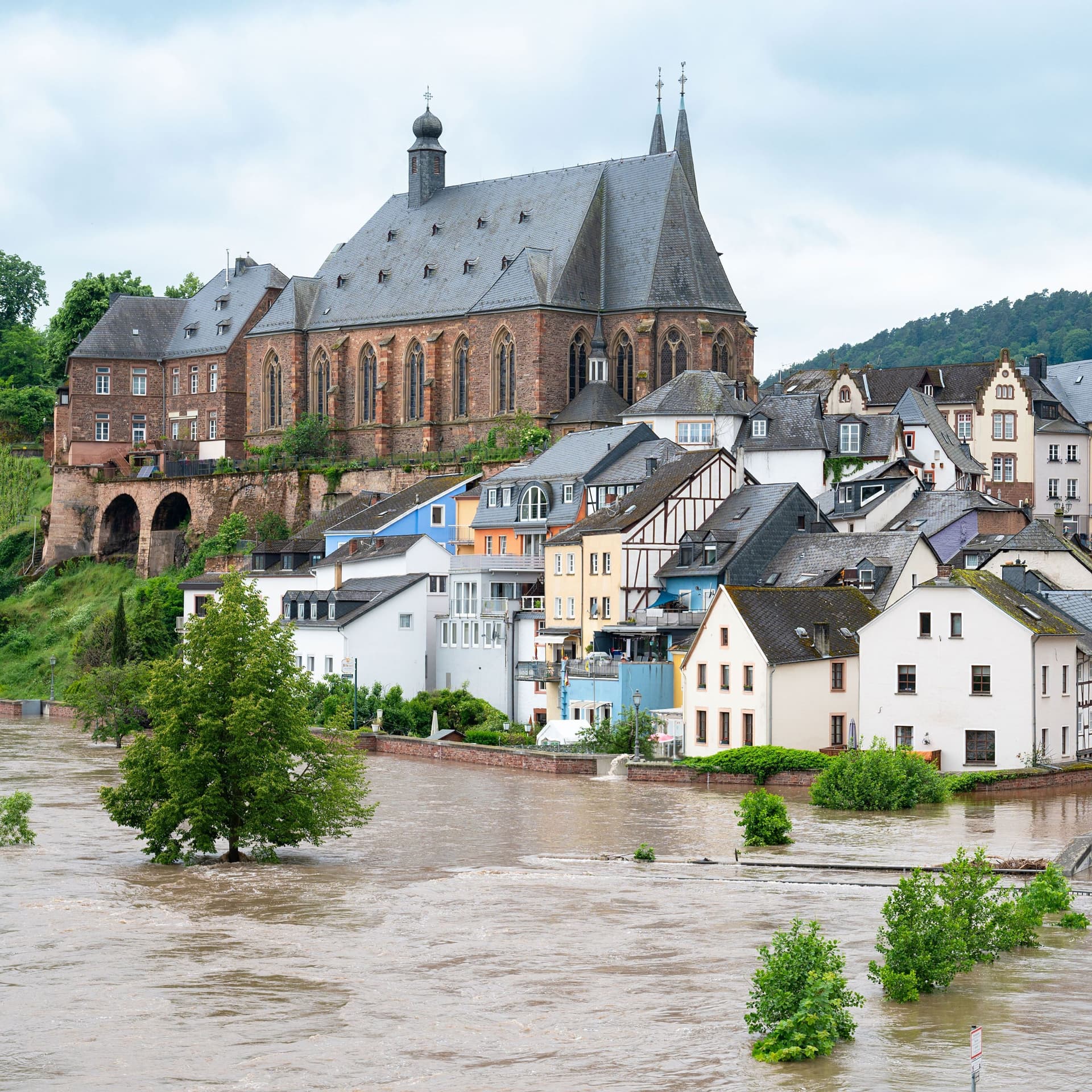Ein überflutete Stadt am Fluss. Auf einem Hügel steht eine Kirche. Davor befinden sich überflutete Wohnhäuser.
