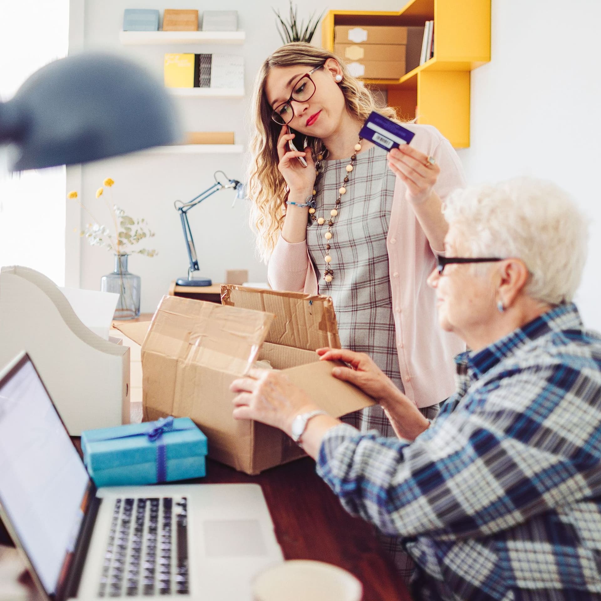 Eine Frau mit Bankkarte in der Hand telefoniert ernst. Eine Seniorin sitzt mit einem geöffneten Paket neben ihr. Ihr Laptop ist aufgeklappt.
