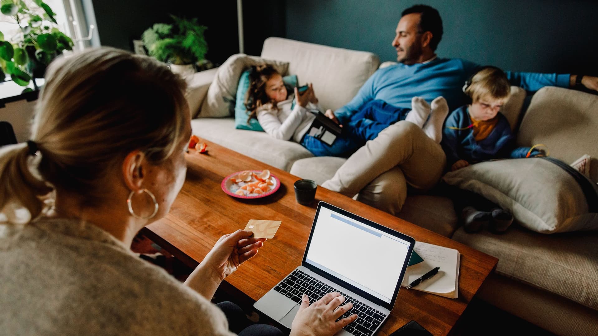 Blick über die Schulter einer Frau, die am Laptop Daten einer Bankkarte eingibt. Im Hintergrund ist ihre Familie auf dem Sofa sitzend.