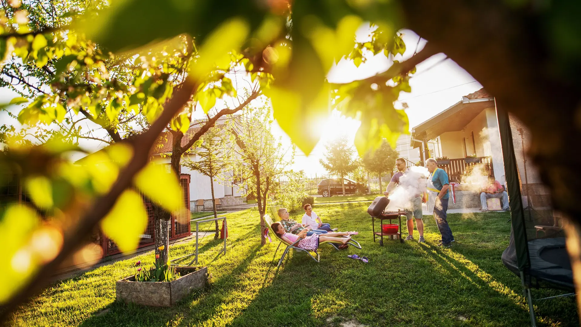 Familie grillt im Garten umgeben von kleineren Bäumen. Die Sonne scheint in die Kamera.