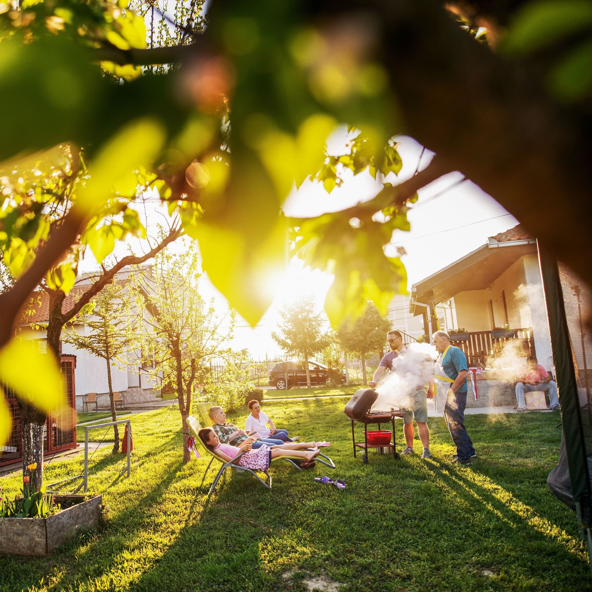 Familie grillt im Garten umgeben von kleineren Bäumen. Die Sonne scheint in die Kamera.