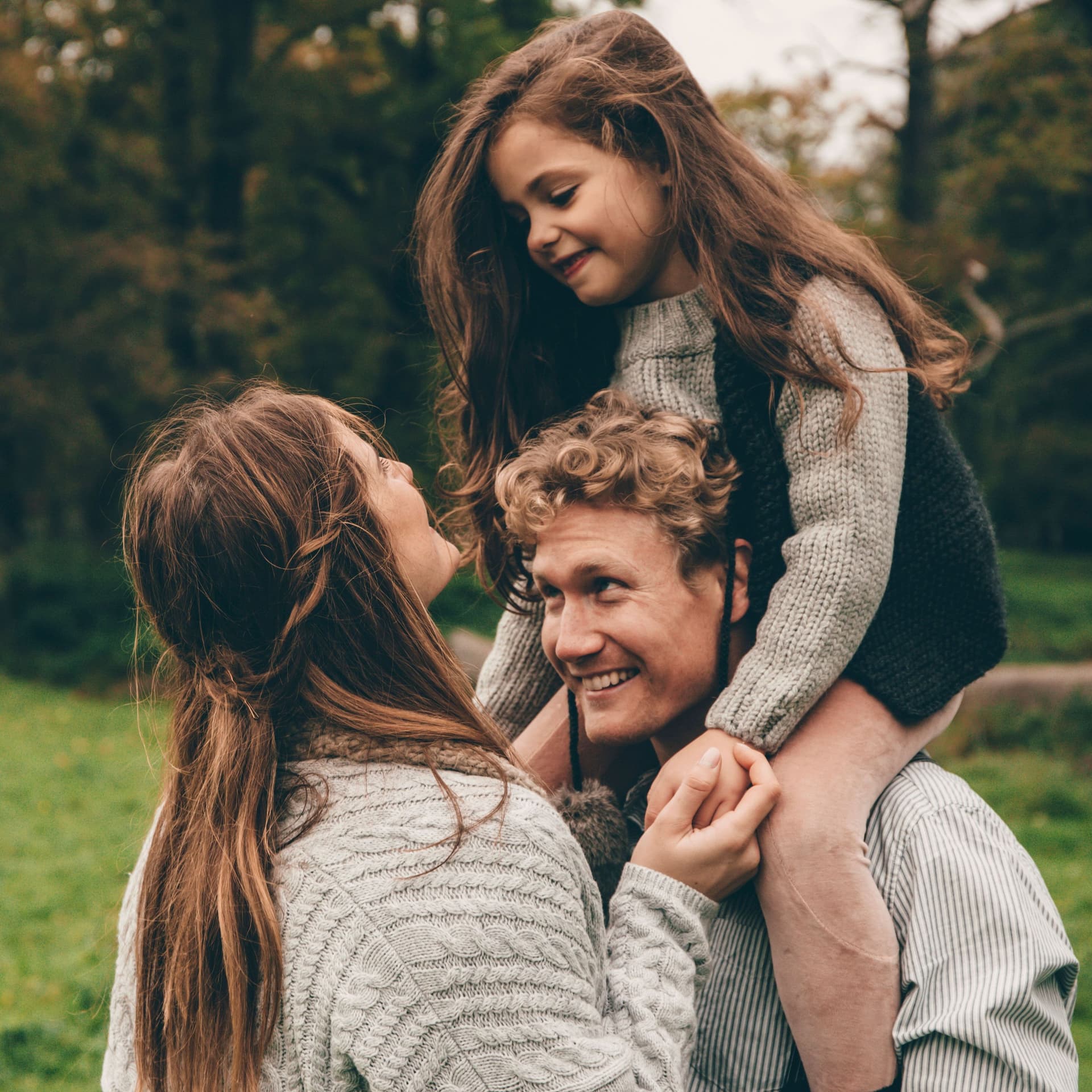 Glückliches Paar mit kleiner Tochter auf den Schultern stehen nah beieinander in der grünen Natur.