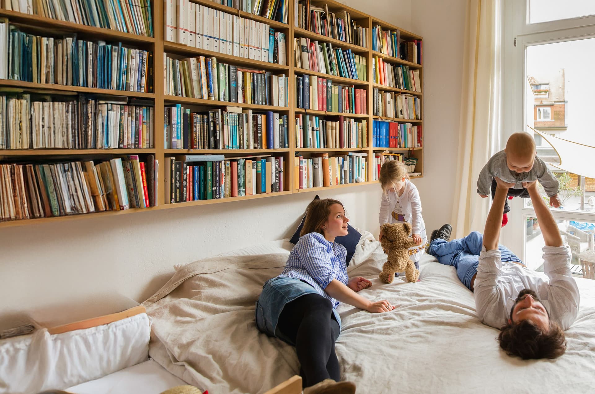 Eine vierköpfige Familie spielt zusammen auf einem Bett. Über dem Bett hängt ein großes Bücherregal.