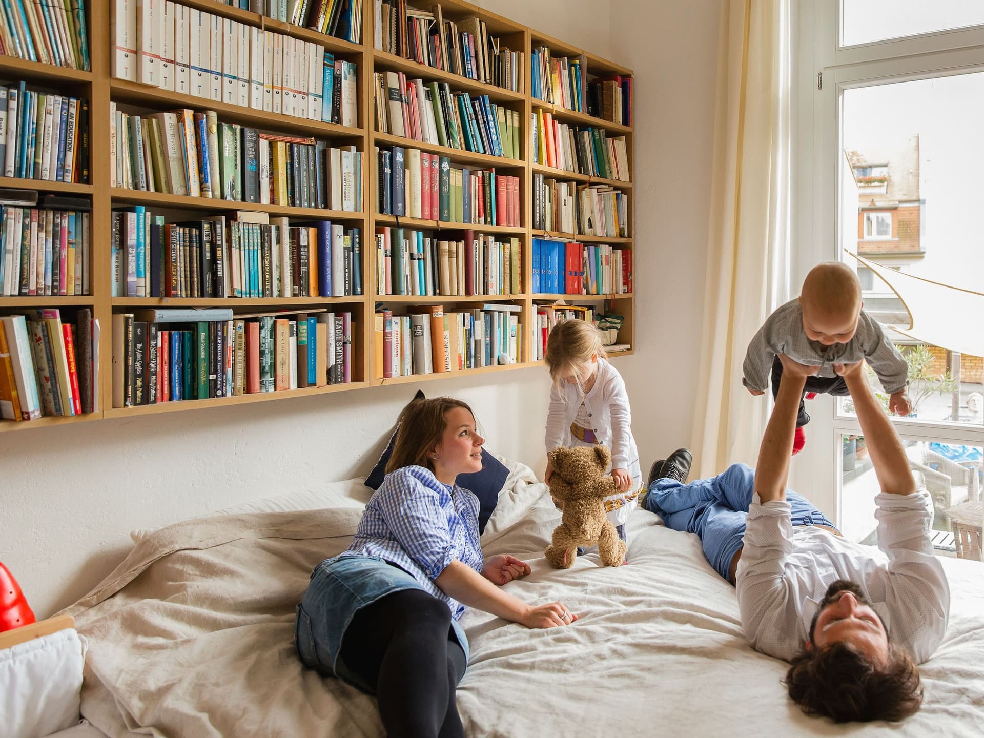 Eine vierköpfige Familie spielt zusammen auf einem Bett. Über dem Bett hängt ein großes Bücherregal.