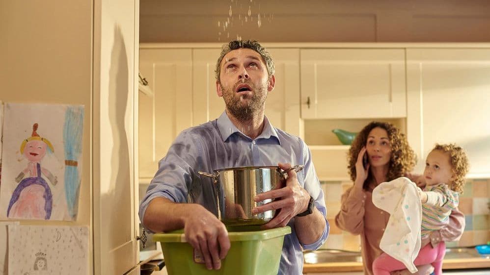 Junge Familie steht in der Küche; es tropft Wasser aus der Zimmerdecke, das der Mann mit einem Topf auffängt.