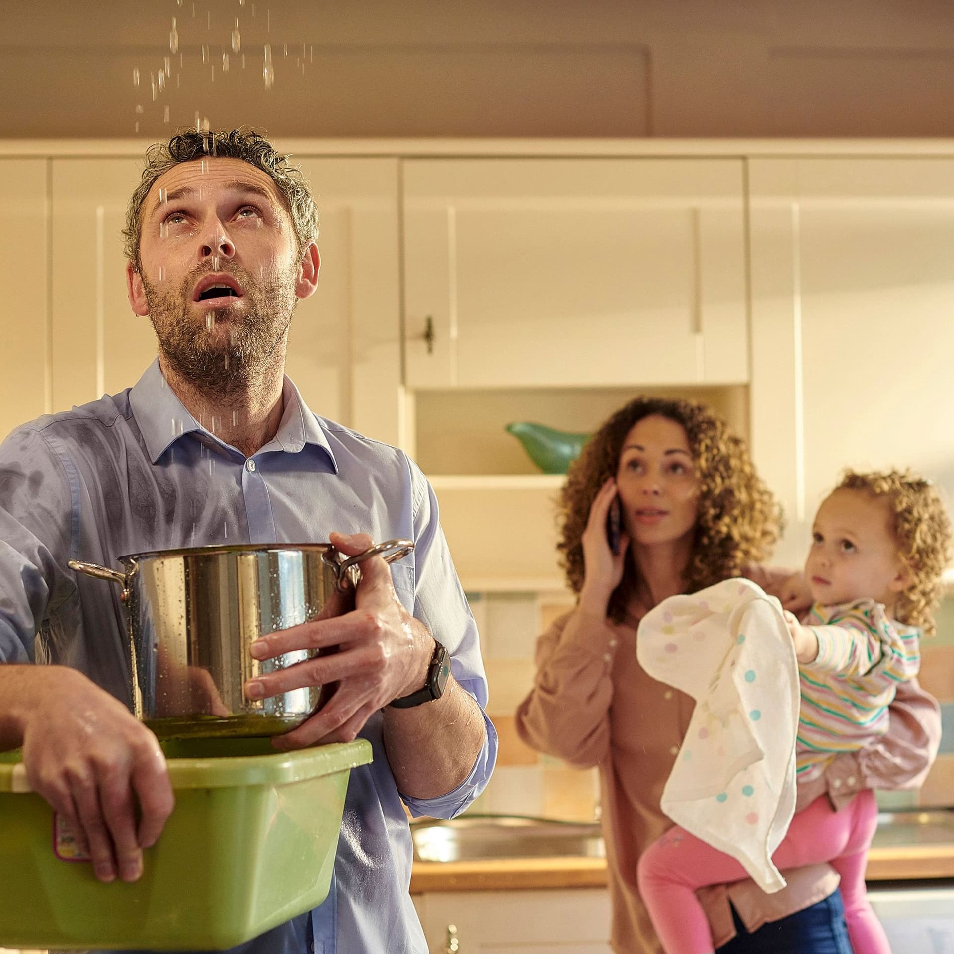 Junge Familie steht in der Küche; es tropft Wasser aus der Zimmerdecke, das der Mann mit einem Topf auffängt.