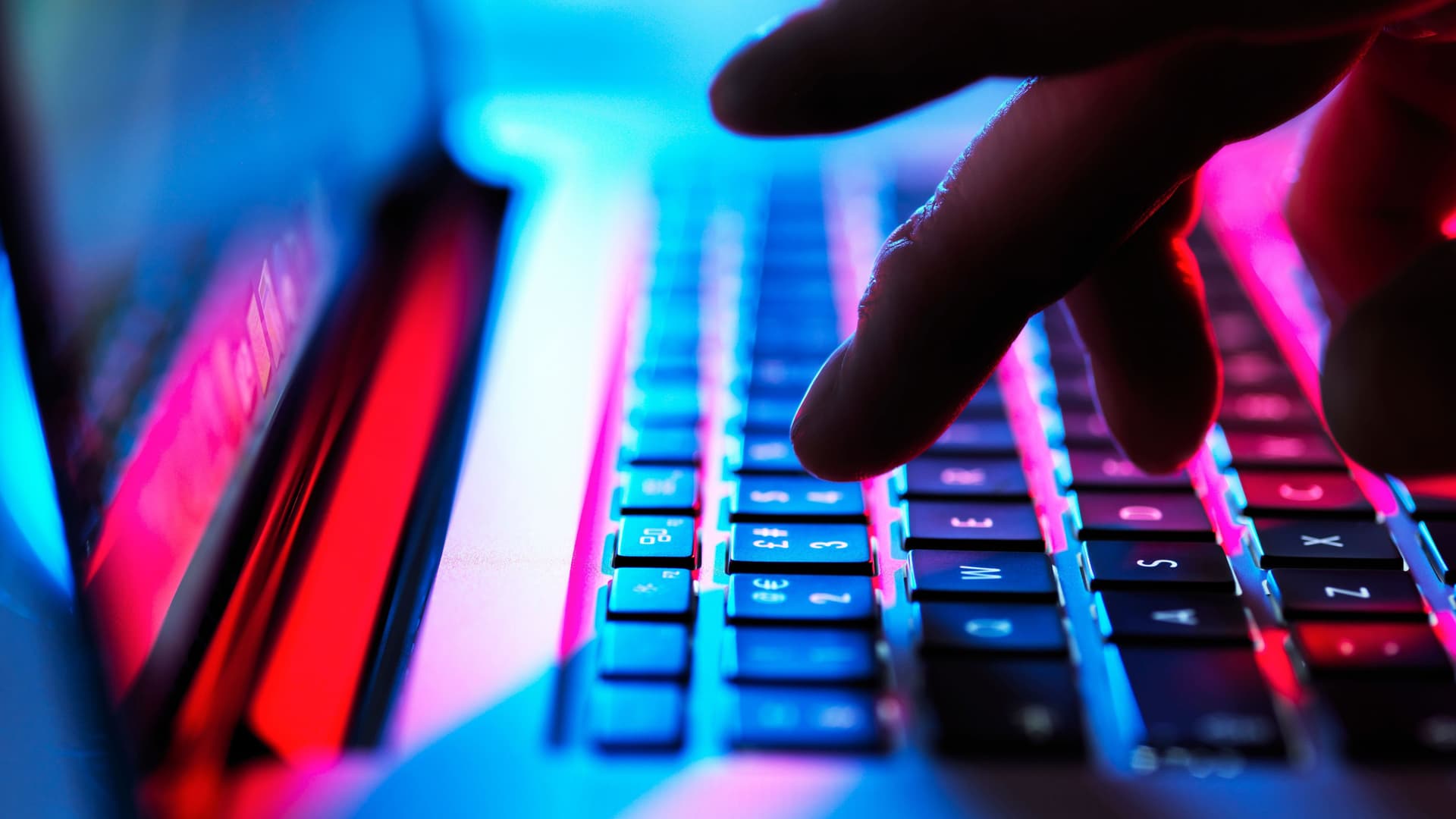 Hand, die im Dunklen auf einer Laptoptastatur tippt. Man sieht blaues und pinkes Licht.