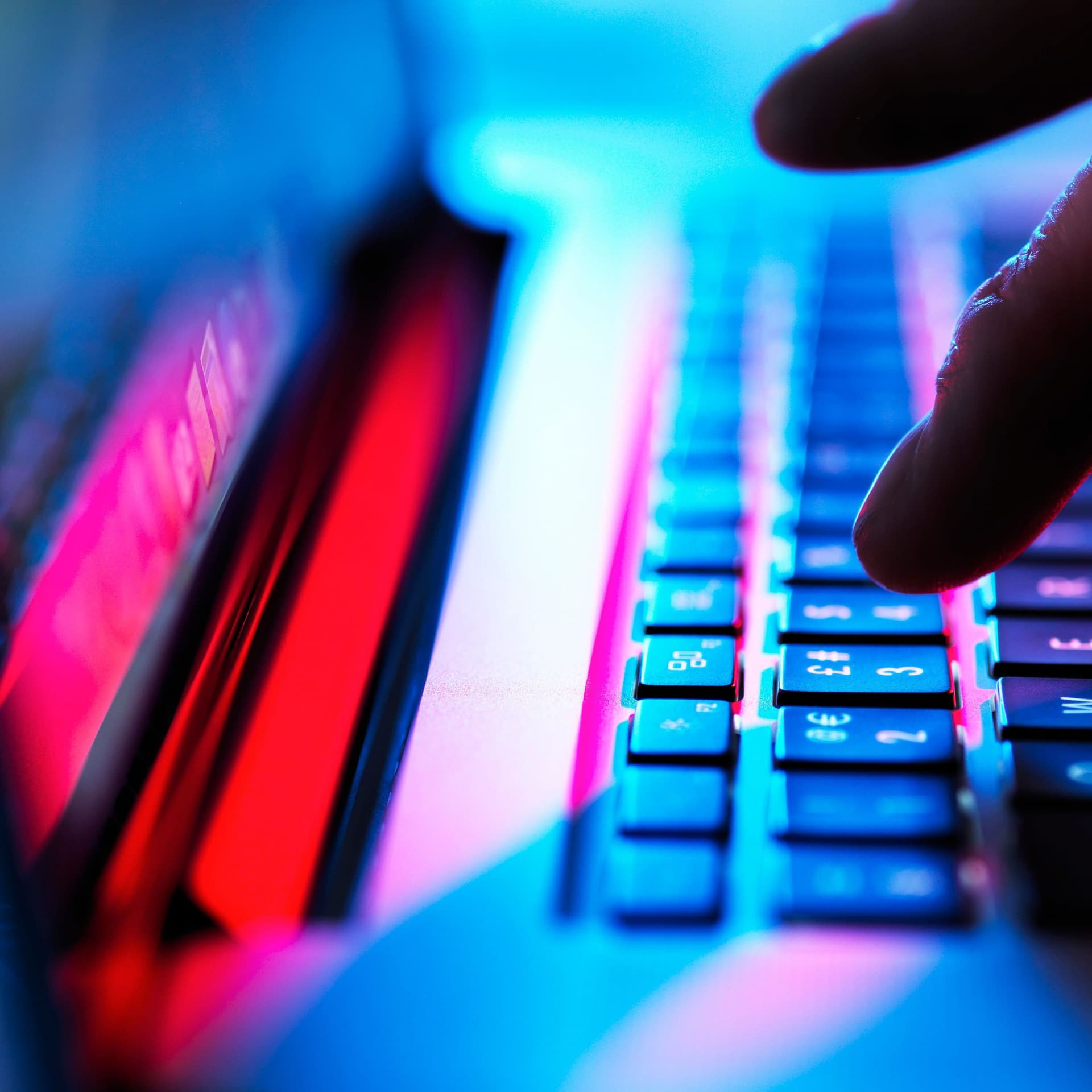 Hand, die im Dunklen auf einer Laptoptastatur tippt. Man sieht blaues und pinkes Licht.