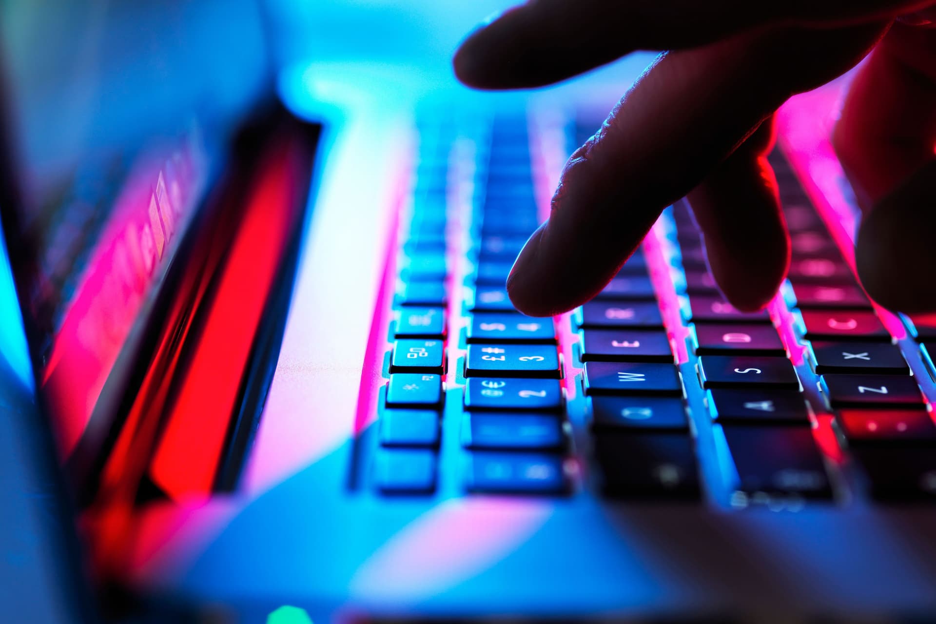 Hand, die im Dunklen auf einer Laptoptastatur tippt. Man sieht blaues und pinkes Licht.