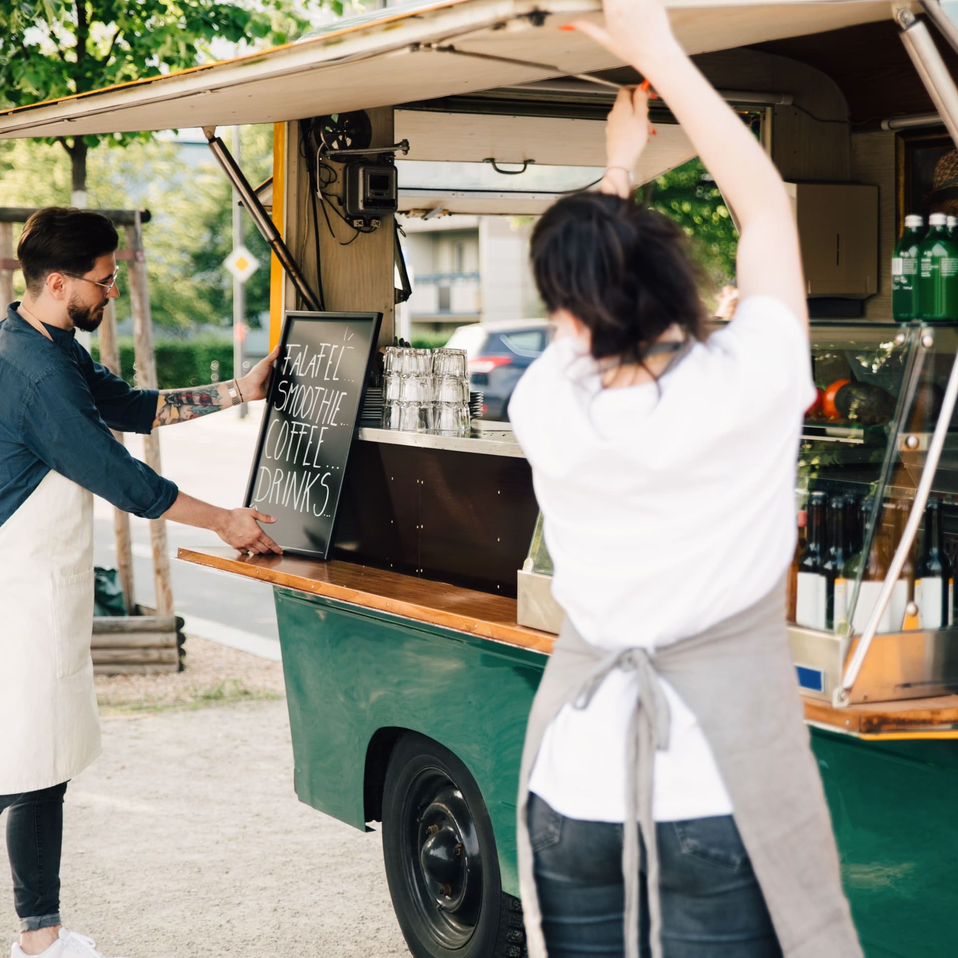 Ein junger Mann mit weißer Schürze passt eine Tafel am Imbisswagen an, während die weibliche Mitarbeiterin mit dem Rücken zur Kamera die Klappe des Imbisswagen öffnet.