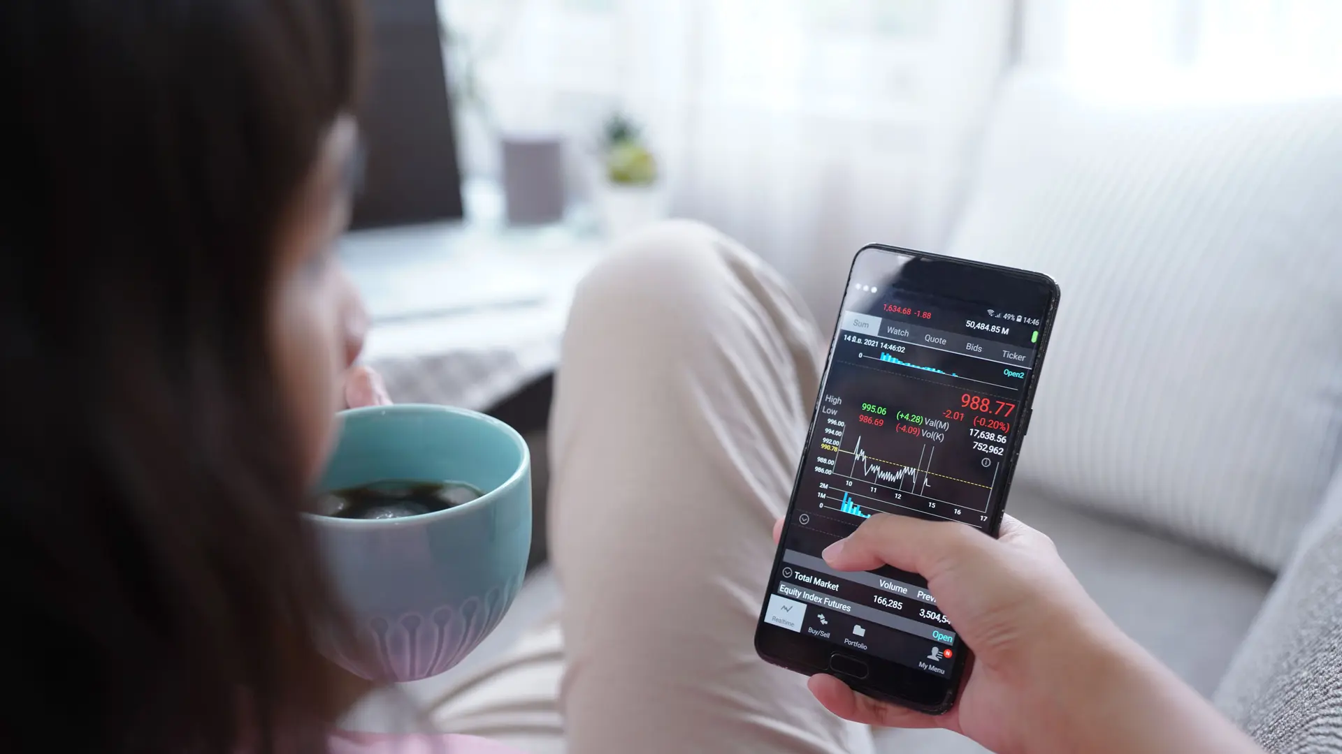 Blick über die Schulter einer Frau mit Kaffee auf einem Sofa. In ihrer Hand hält sie ihr Smartphone, auf dem sie mobil per App investiert.
