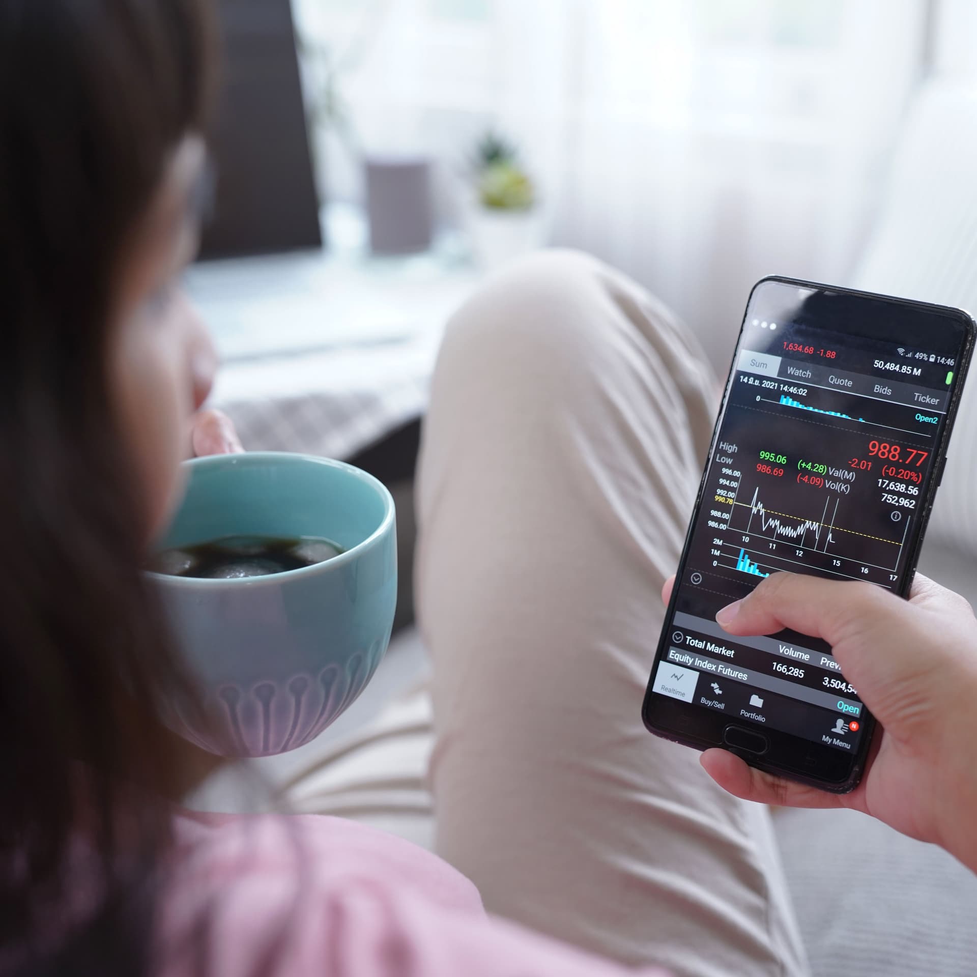 Blick über die Schulter einer Frau mit Kaffee auf einem Sofa. In ihrer Hand hält sie ihr Smartphone, auf dem sie mobil per App investiert.
