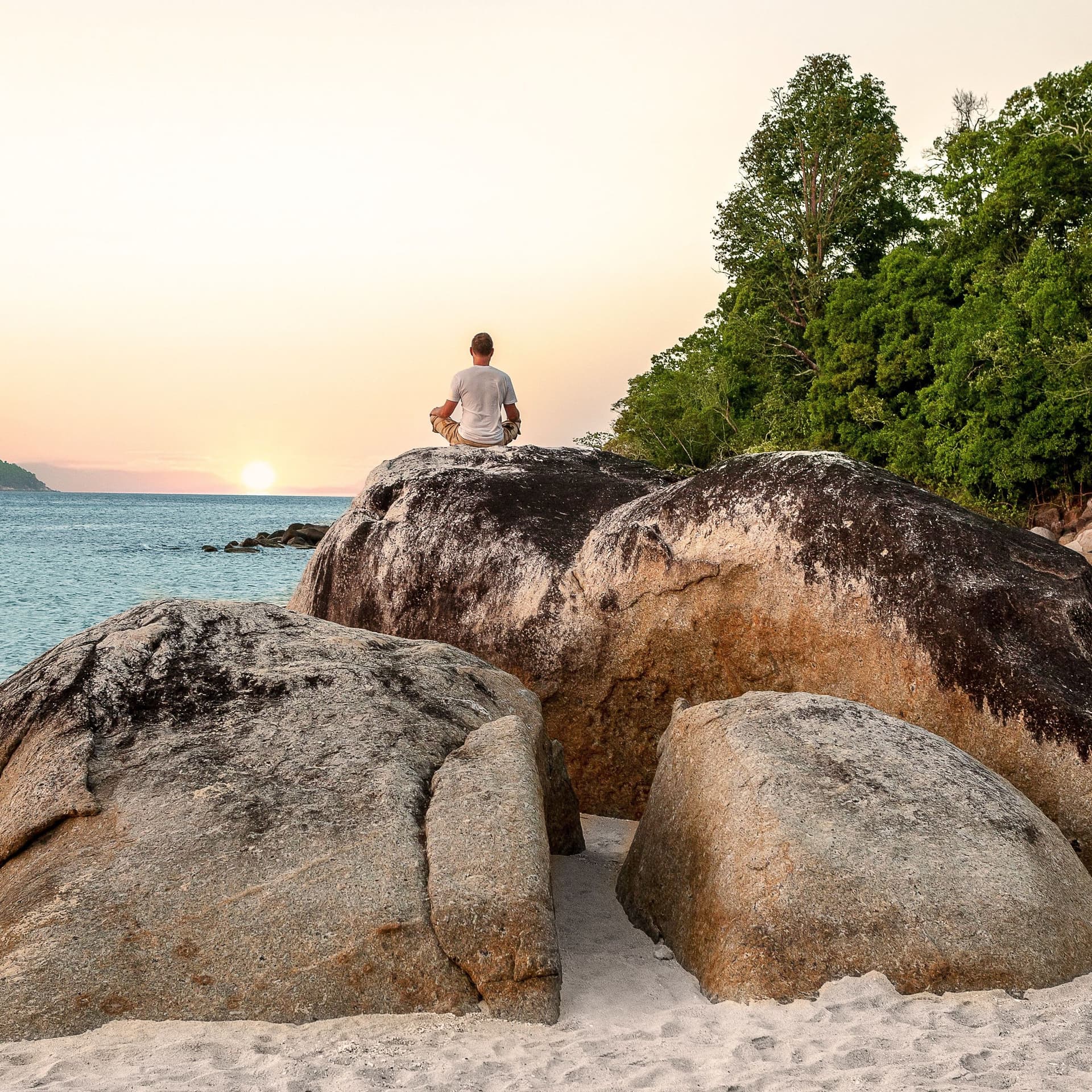 Mann sitzt im Schneidersitz auf Felsen am Strand