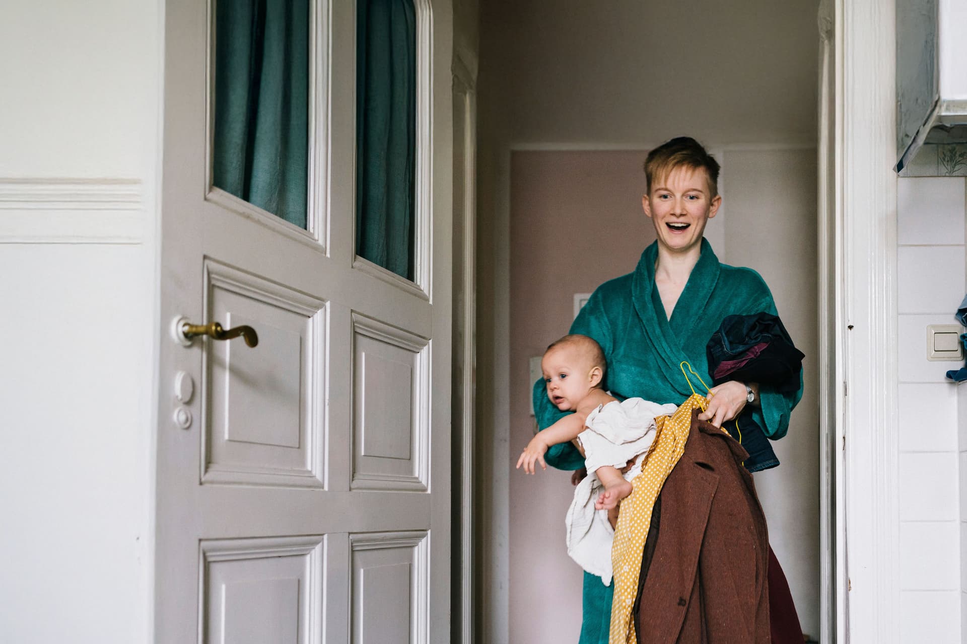Eine junge Frau, die einige Kleider und ein Baby in ein Badezimmer trägt. Sie trägt einen Bademantel und lächelt mit geöffnetem Mund.