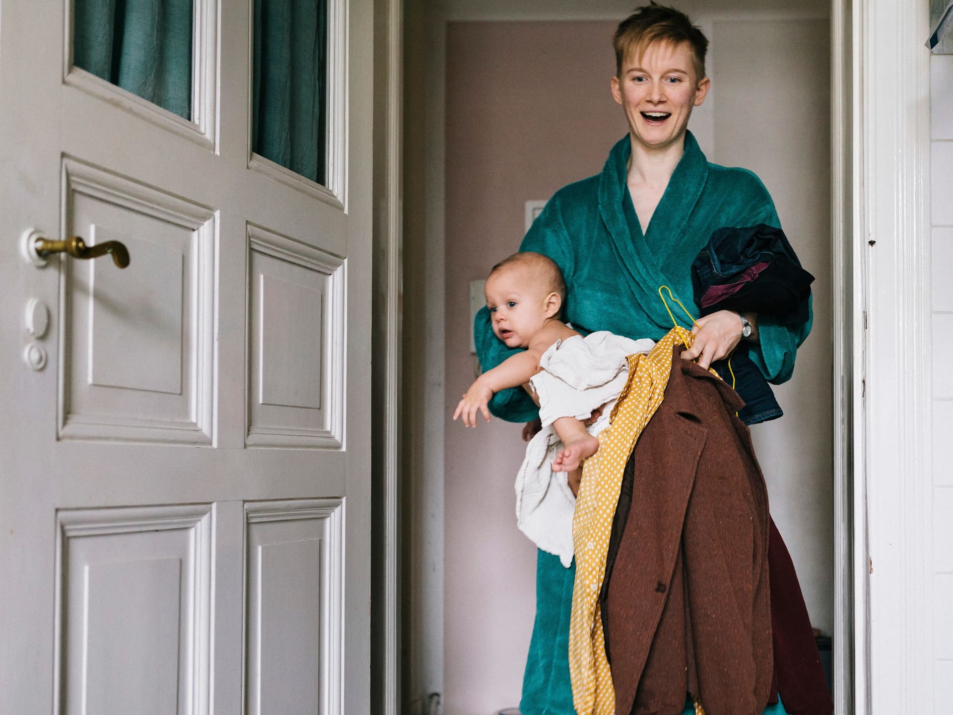 Eine junge Frau, die einige Kleider und ein Baby in ein Badezimmer trägt. Sie trägt einen Bademantel und lächelt mit geöffnetem Mund.