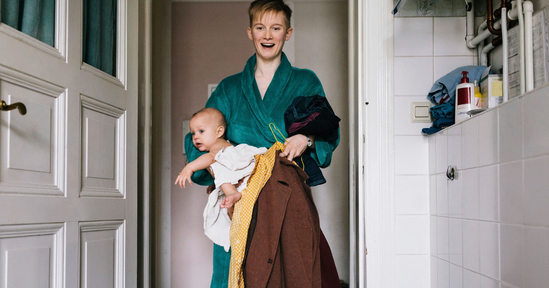 Eine junge Frau, die einige Kleider und ein Baby in ein Badezimmer trägt. Sie trägt einen Bademantel und lächelt mit geöffnetem Mund.