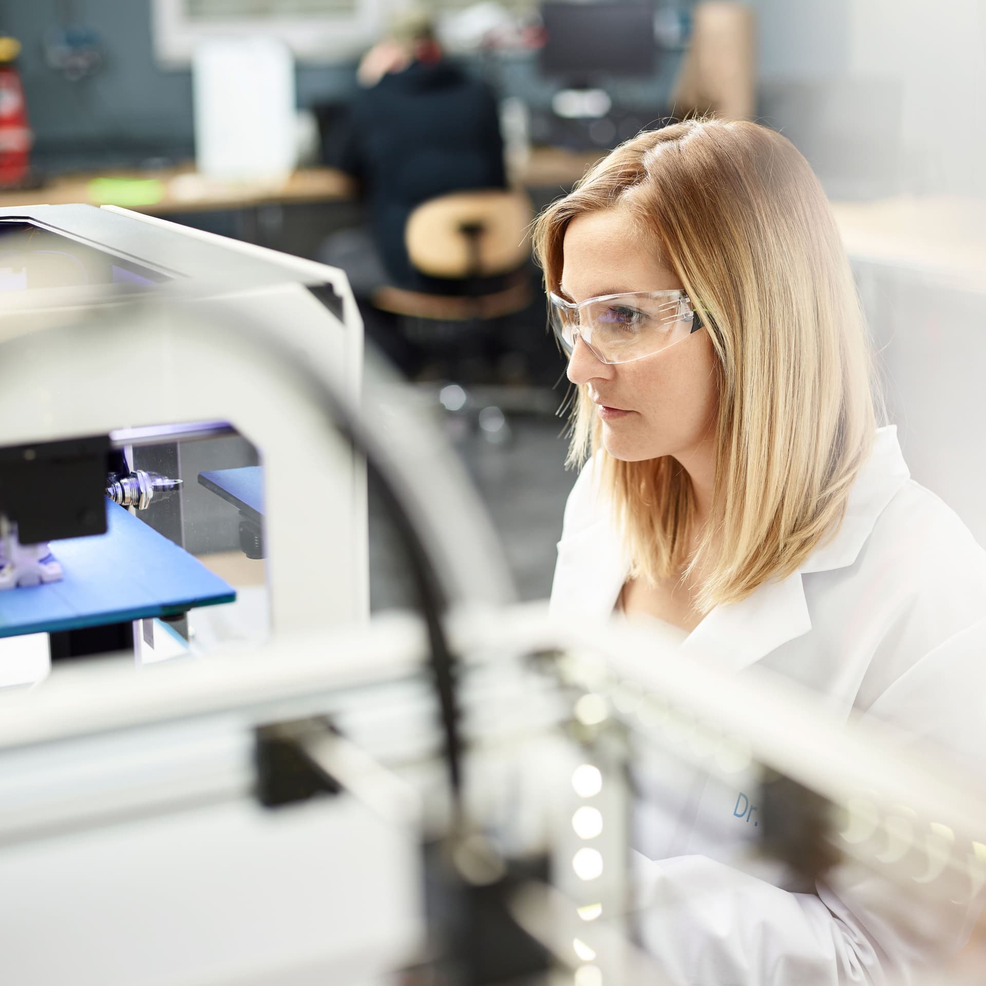 Junge Frau mit Schutzbrille und weißem Kittel sitzt vor einem 3D Printer und bedient diesen konzentriert
