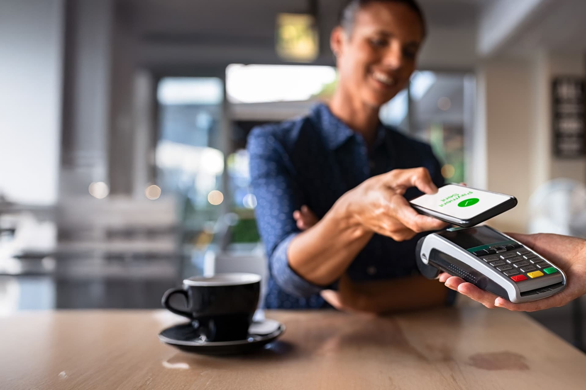 Eine Frau bezahlt in einem Café mit ihrem Smartphone. Dabei hält sie ihr Smartphone vor ein Kartenlesegerät.