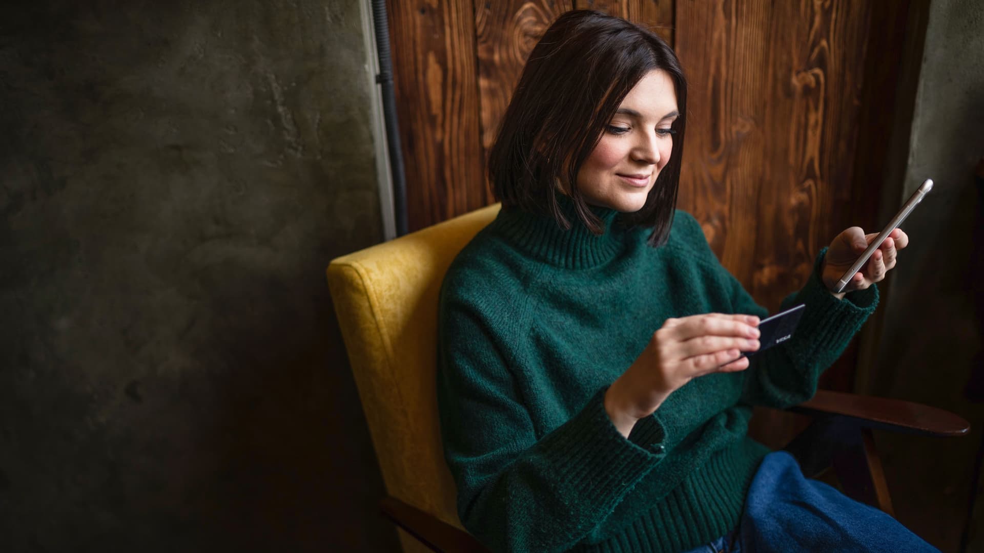 Lächelnde Frau, die online mit einer Kreditkarte und einem Smartphone im Wohnzimmer einkauft.