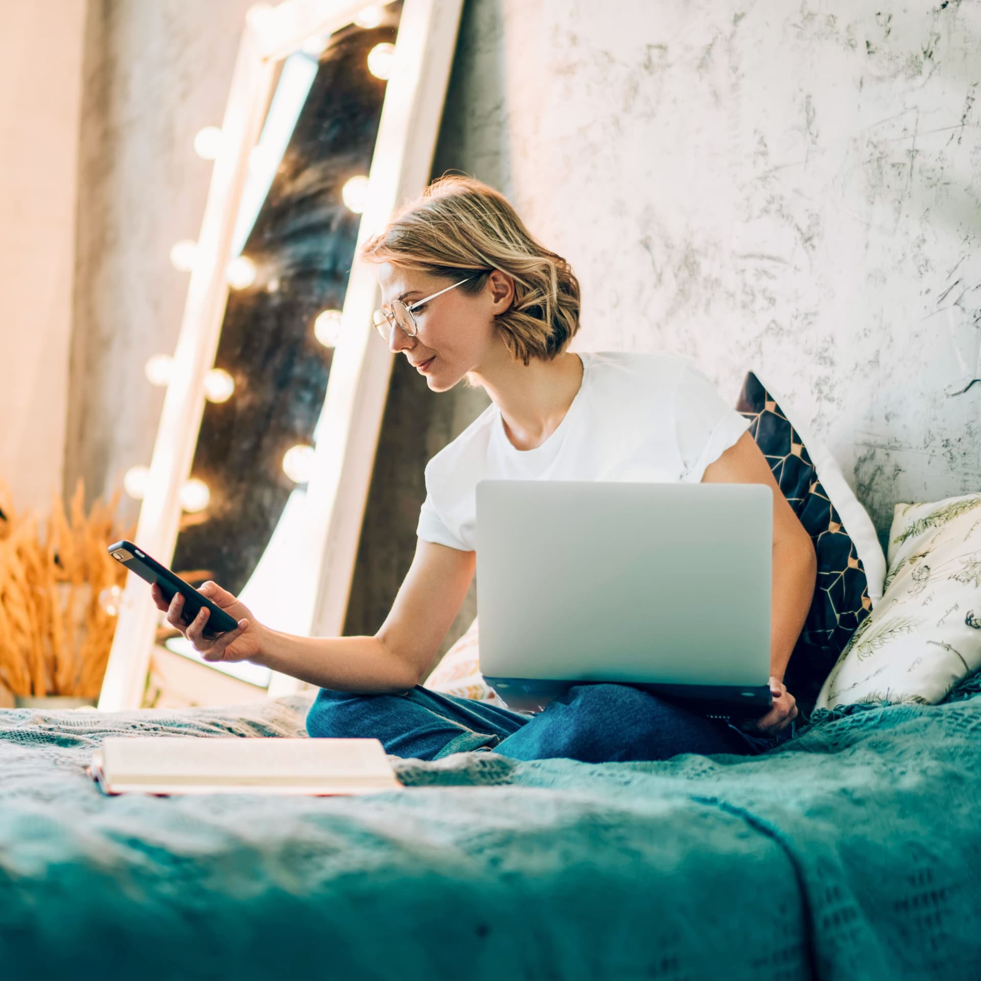 Frau sitzt im Schneidersitz auf dem Bett im Schlafzimmer und schaut auf ihr Handy. Auf dem Bein steht ein Laptop.