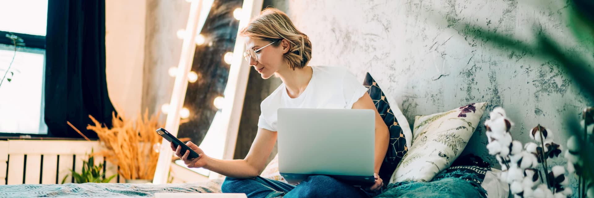 Frau sitzt im Schneidersitz auf dem Bett im Schlafzimmer und schaut auf ihr Handy. Auf dem Bein steht ein Laptop.