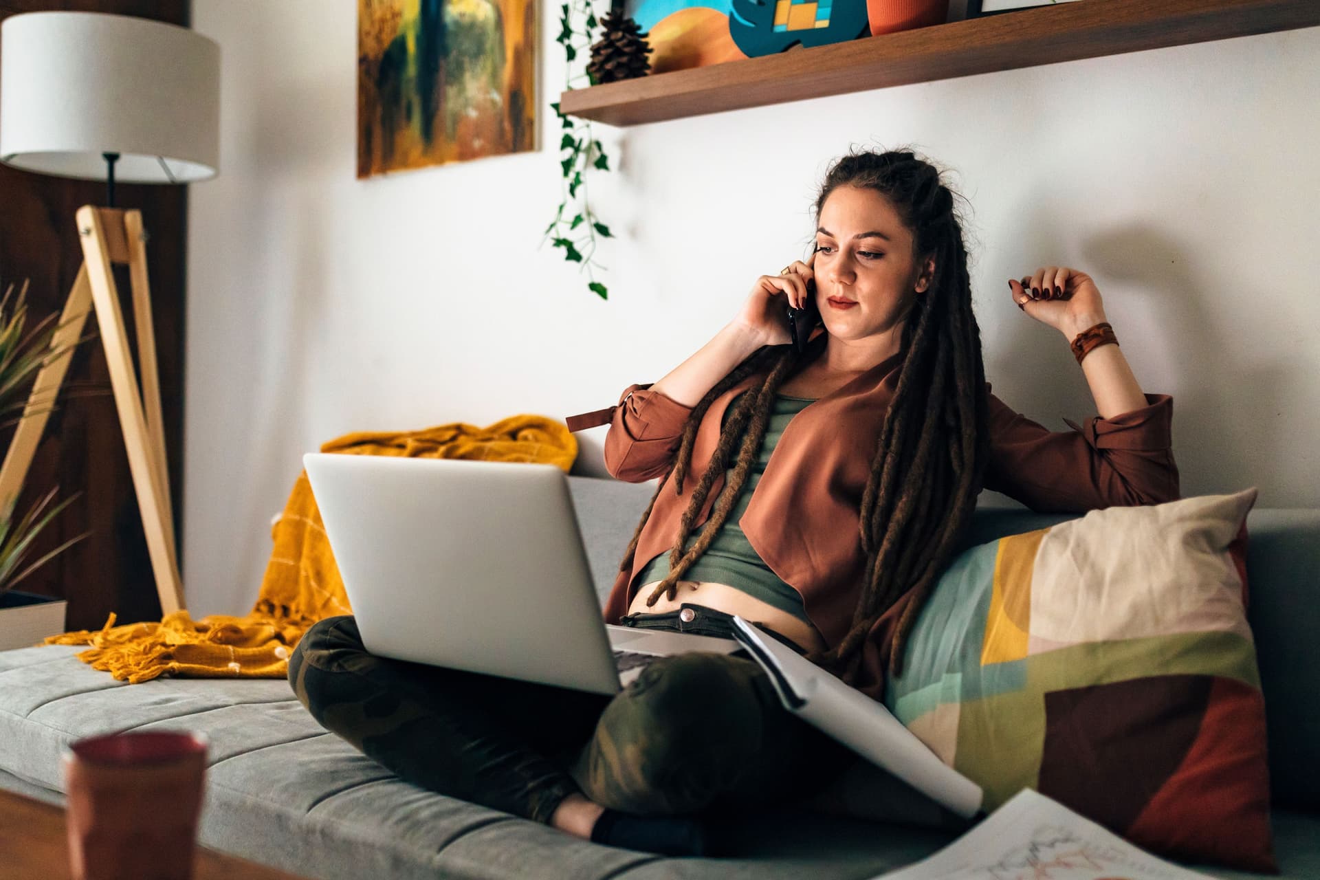 Frau mit langen Dreadlocks arbeitet im Schneidersitz auf dem Sofa. Ihr Laptop steht auf dem Schoß währen sie dabei telefoniert.