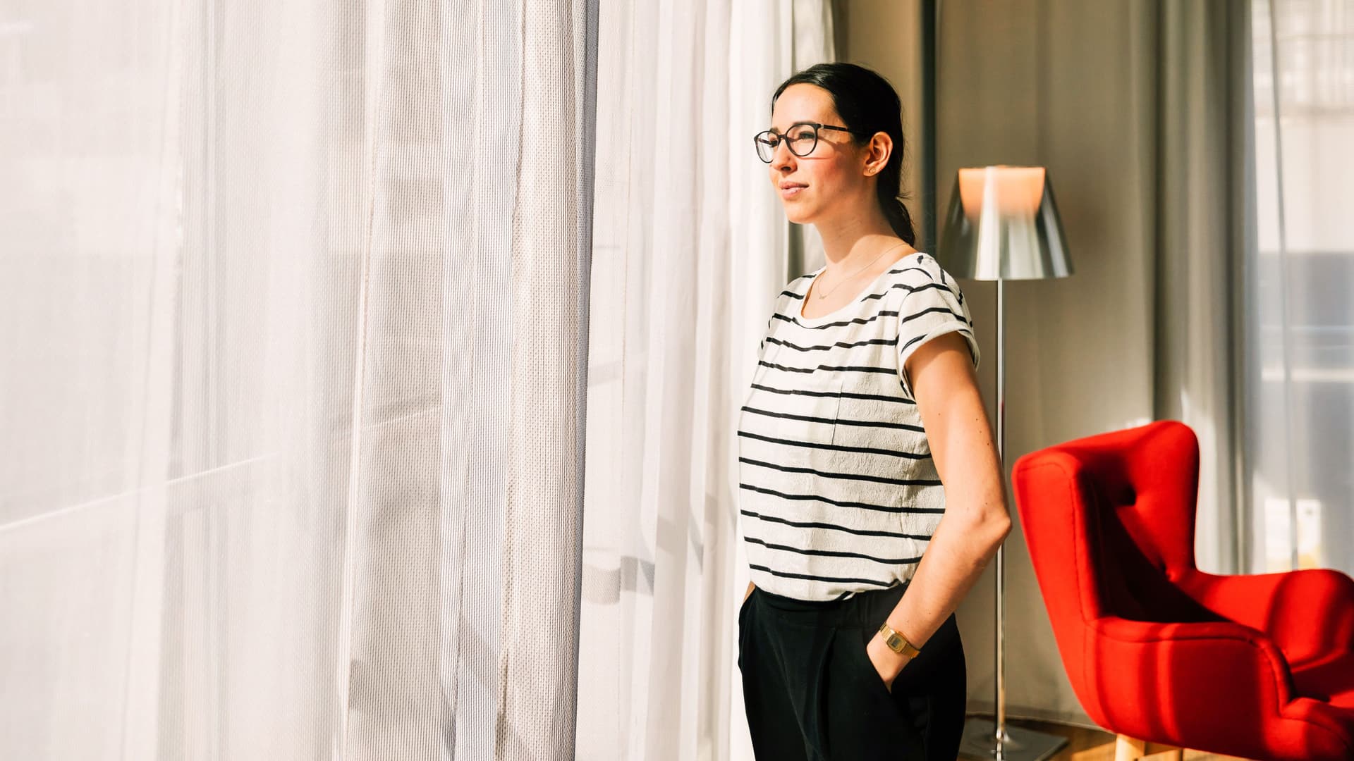 Eine junge Frau steht am Fenster eines hellen, modernen Hotelzimmers.