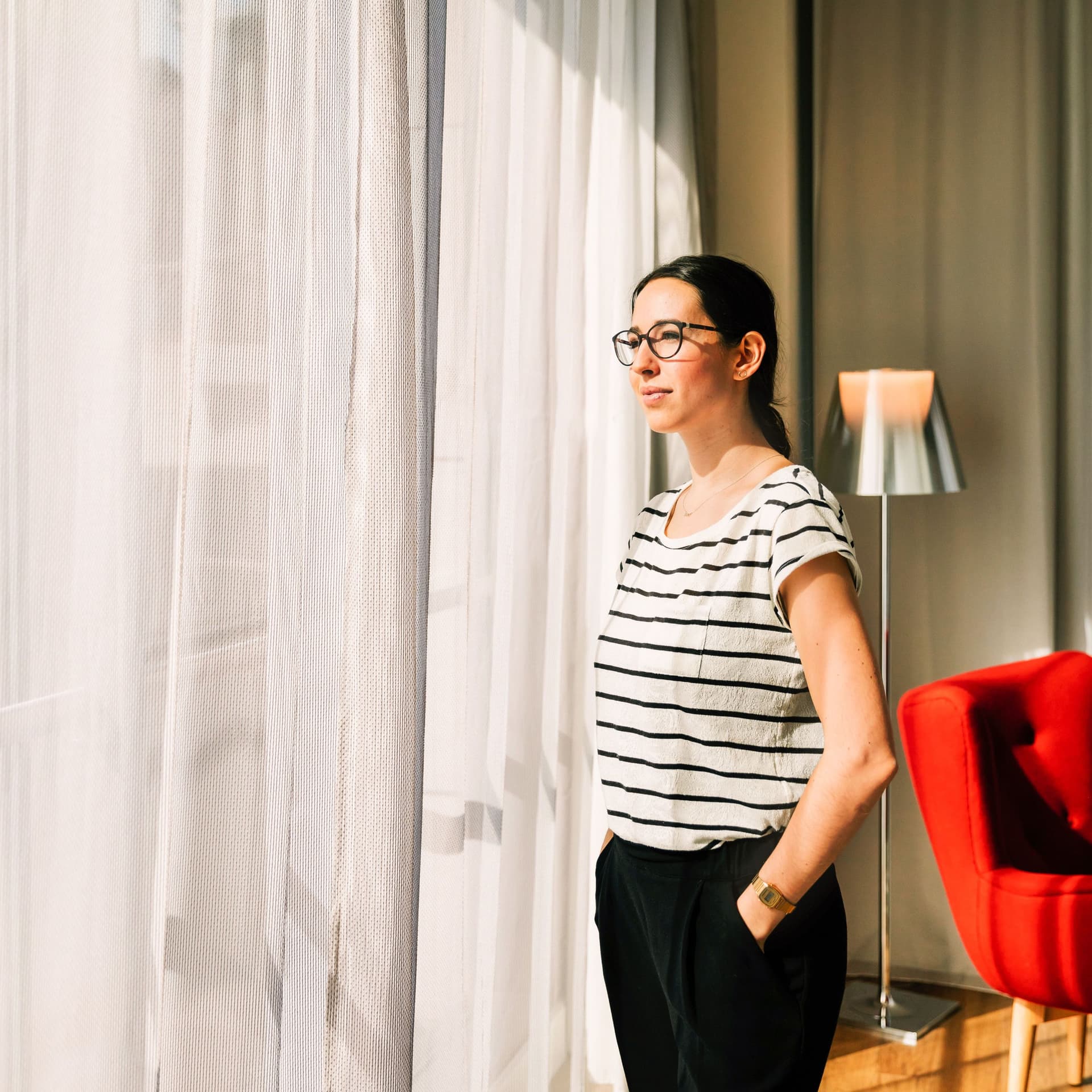 Eine junge Frau steht am Fenster eines hellen, modernen Hotelzimmers.