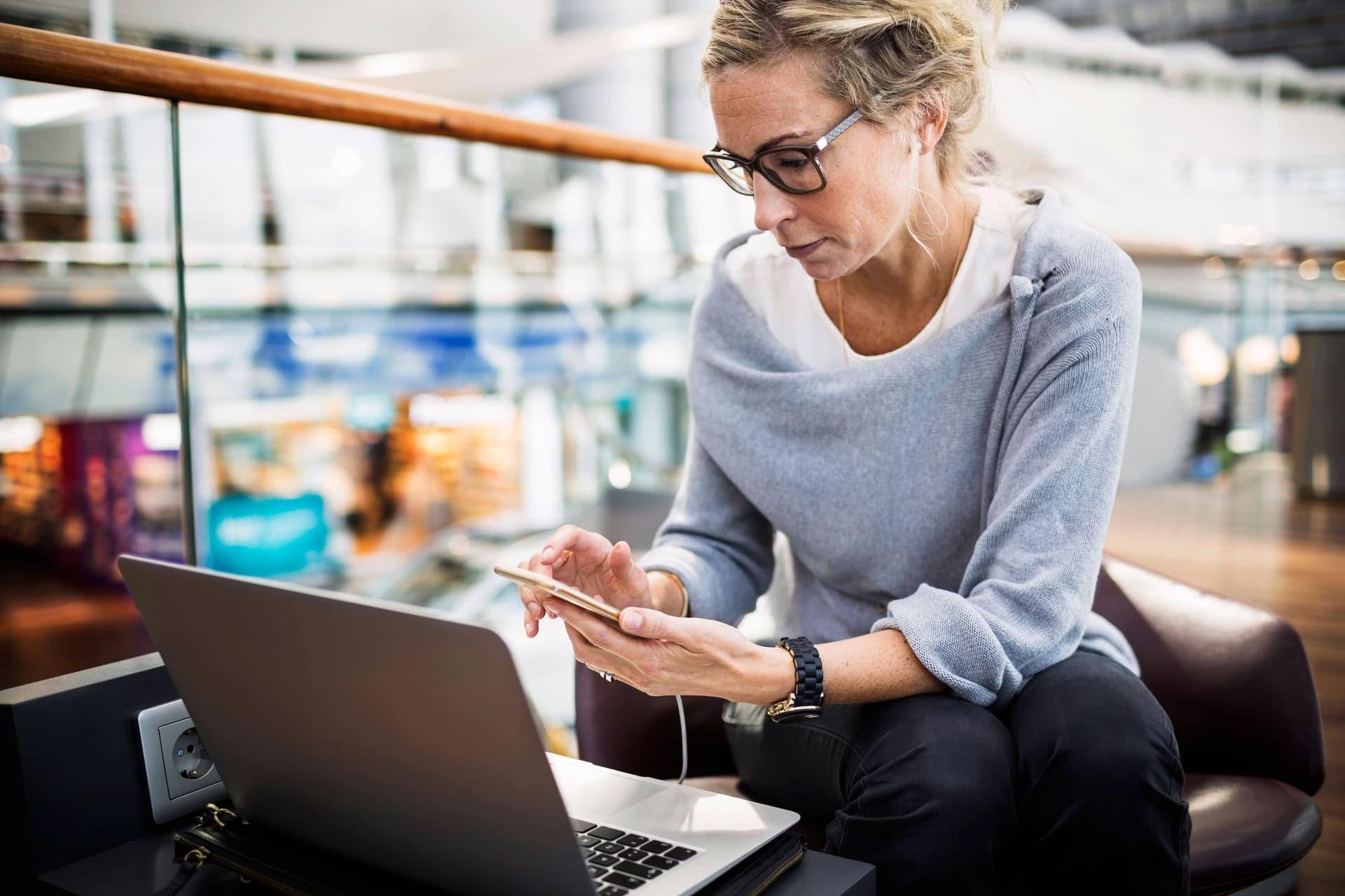 Ein Frau sitzt mit Smartphone und Laptop in einer Flughafenlobby.