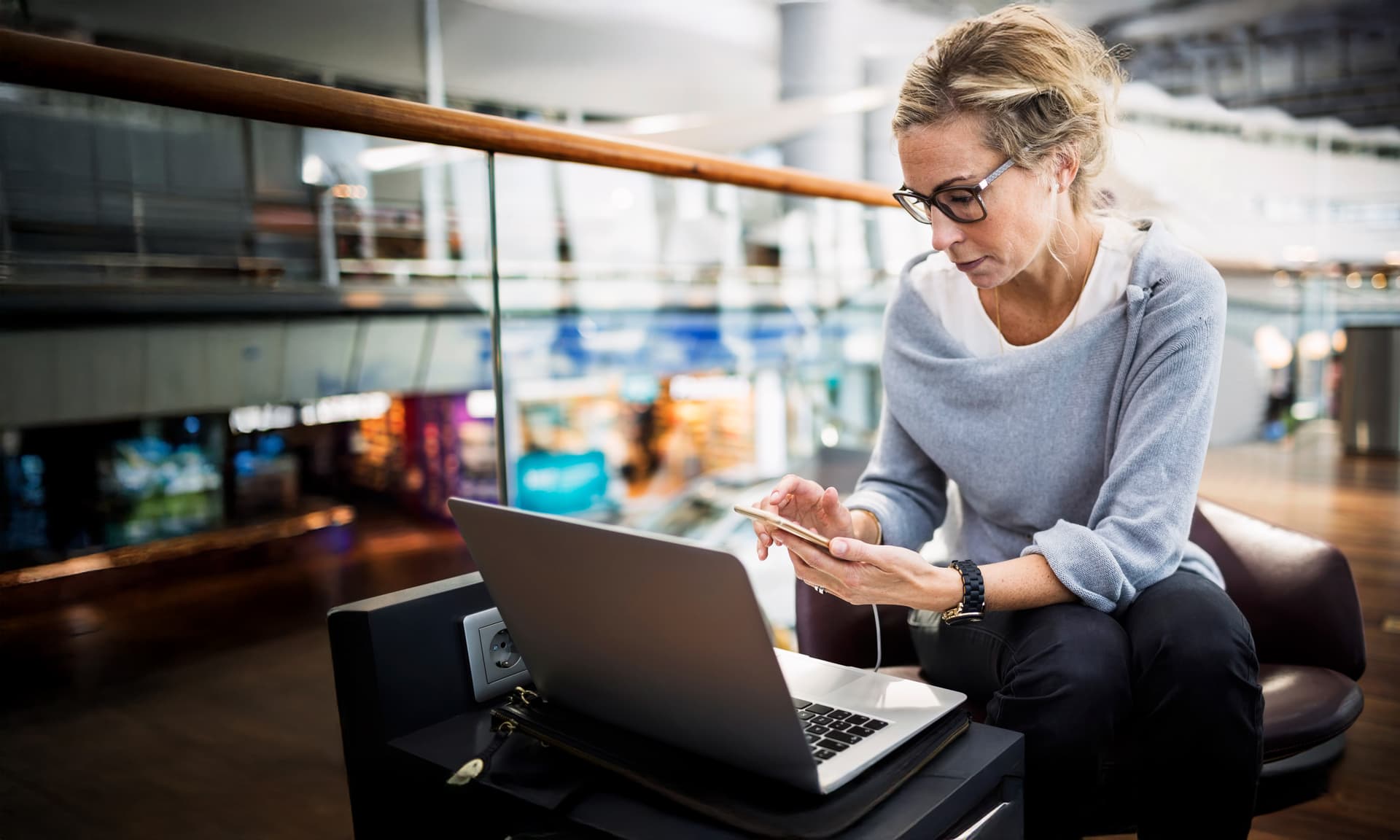 Ein Frau sitzt mit Smartphone und Laptop in einer Flughafenlobby.