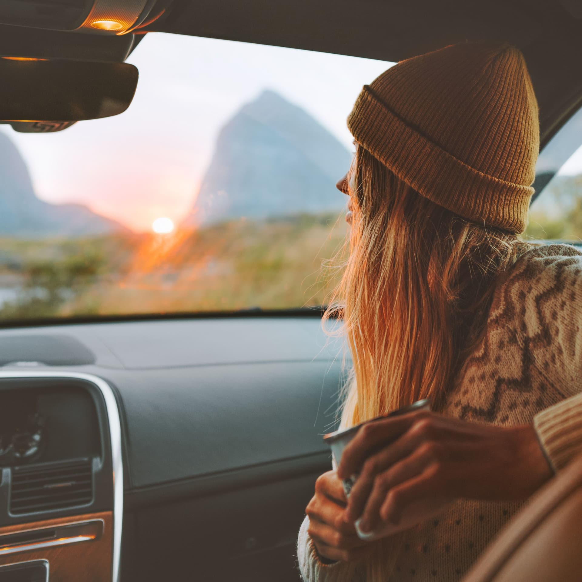 Junge Frau sitzt im Auto und durch die Frontscheibe. Im Hintergrund sind Berge und ein Sonnenuntergang zu sehen.
