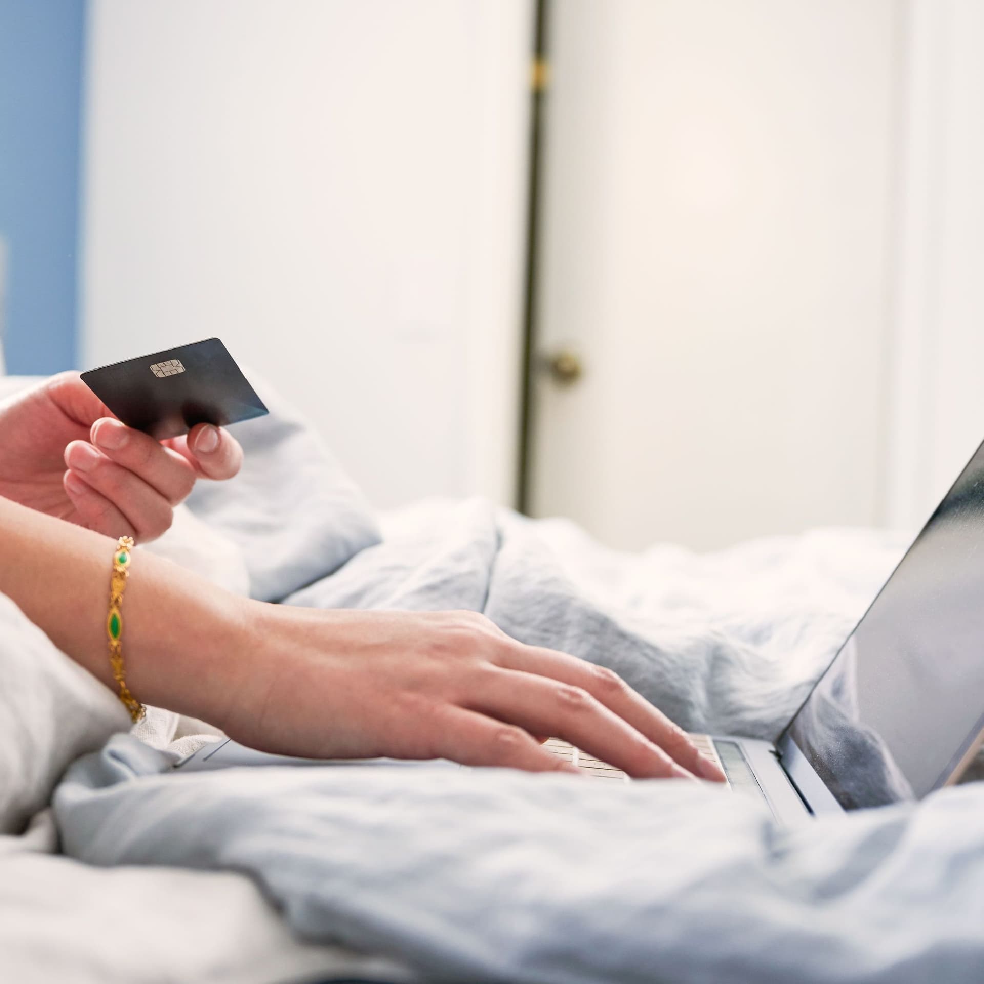 Nahaufnahme einer Frau im Bett sitzend mit einer Bankkarte in der Hand. Mit der anderen bedient sie einen Laptop.