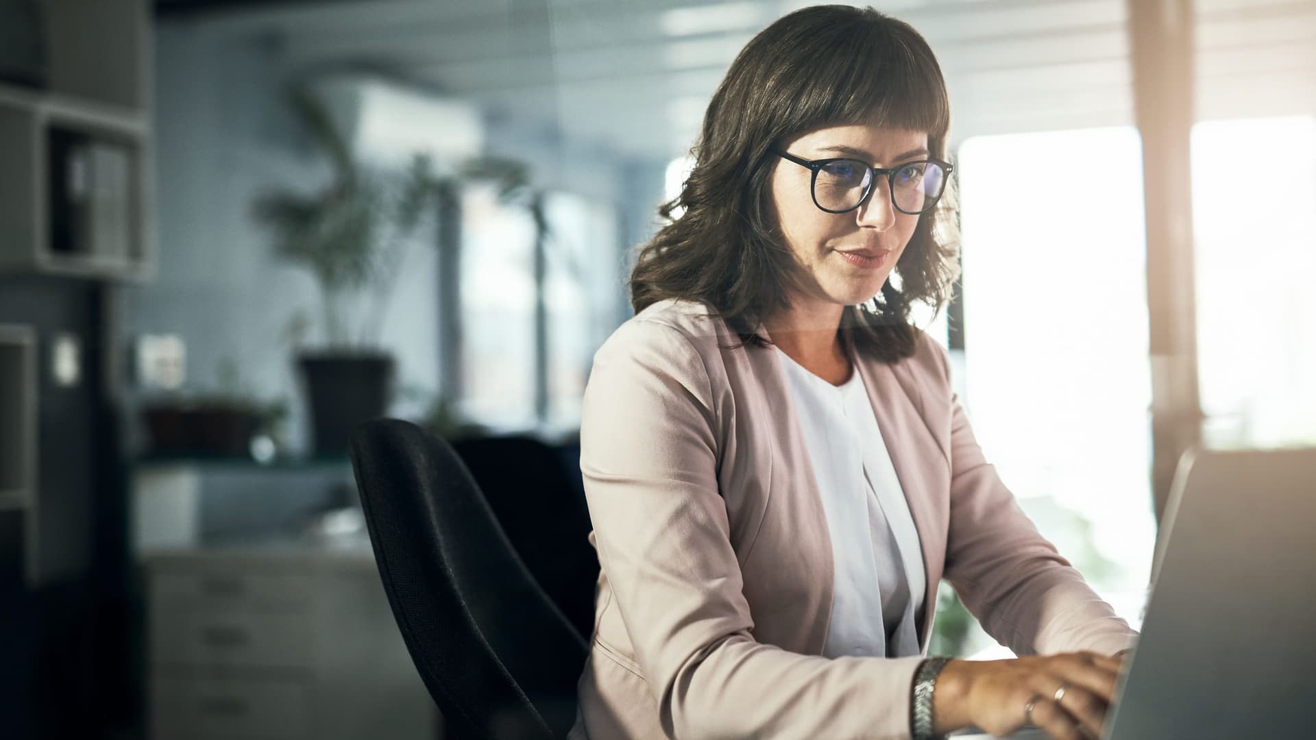 Seitenansicht einer Frau mit Brille mittleren Alters, die in einer abendlichen Lichtstimmung alleine vor einem Laptop sitzt. Im Hintergrund ist ein modernes Büro erkennbar.