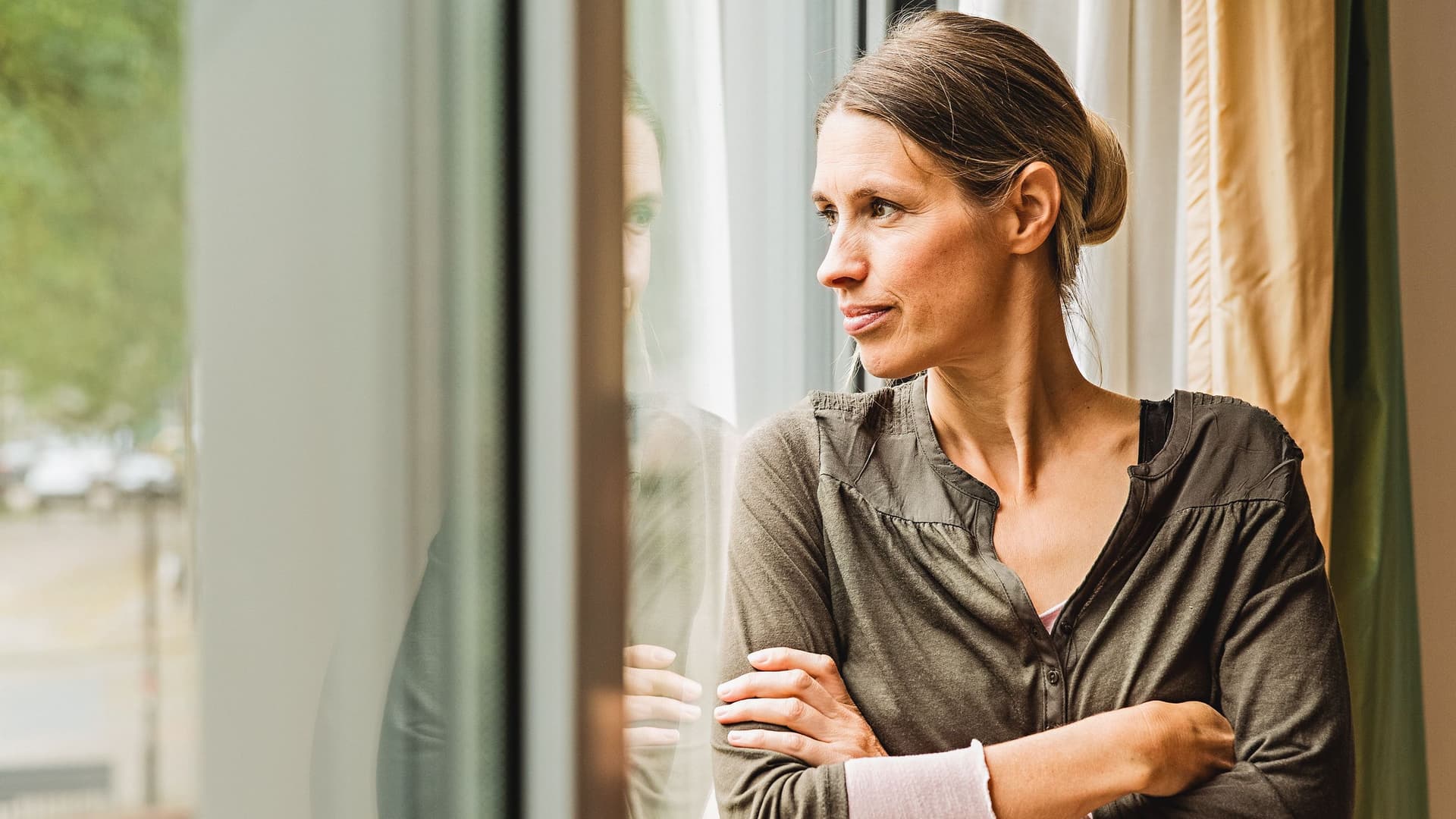Frau mittleren Alters steht gedankenversunken am Fenster