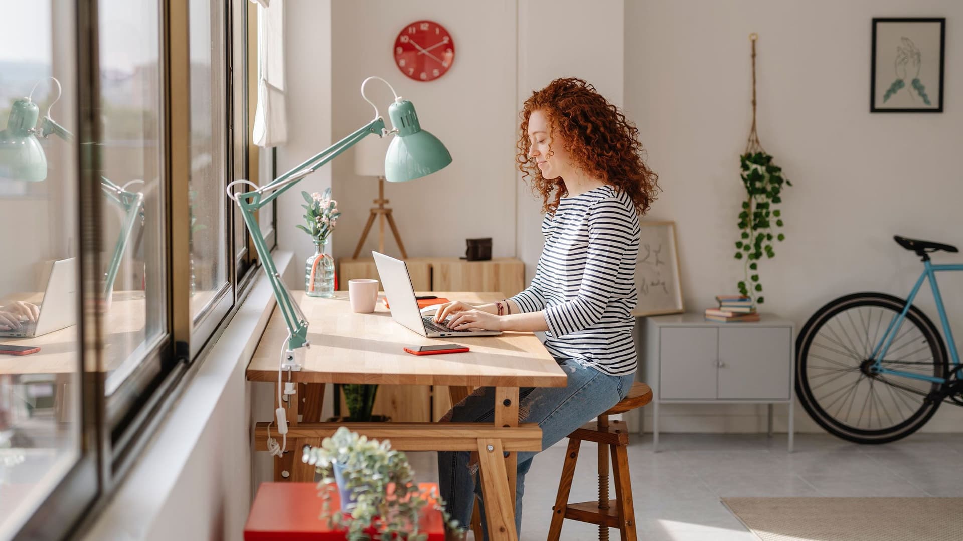Eine junge Frau mit roten Locken arbeitet konzentriert an ihrem Schreibtisch in einem hellen, gemütlichen Homeoffice. Das Zimmer ist stilvoll eingerichtet mit modernen Möbeln, Pflanzen und dekorativen Elementen.