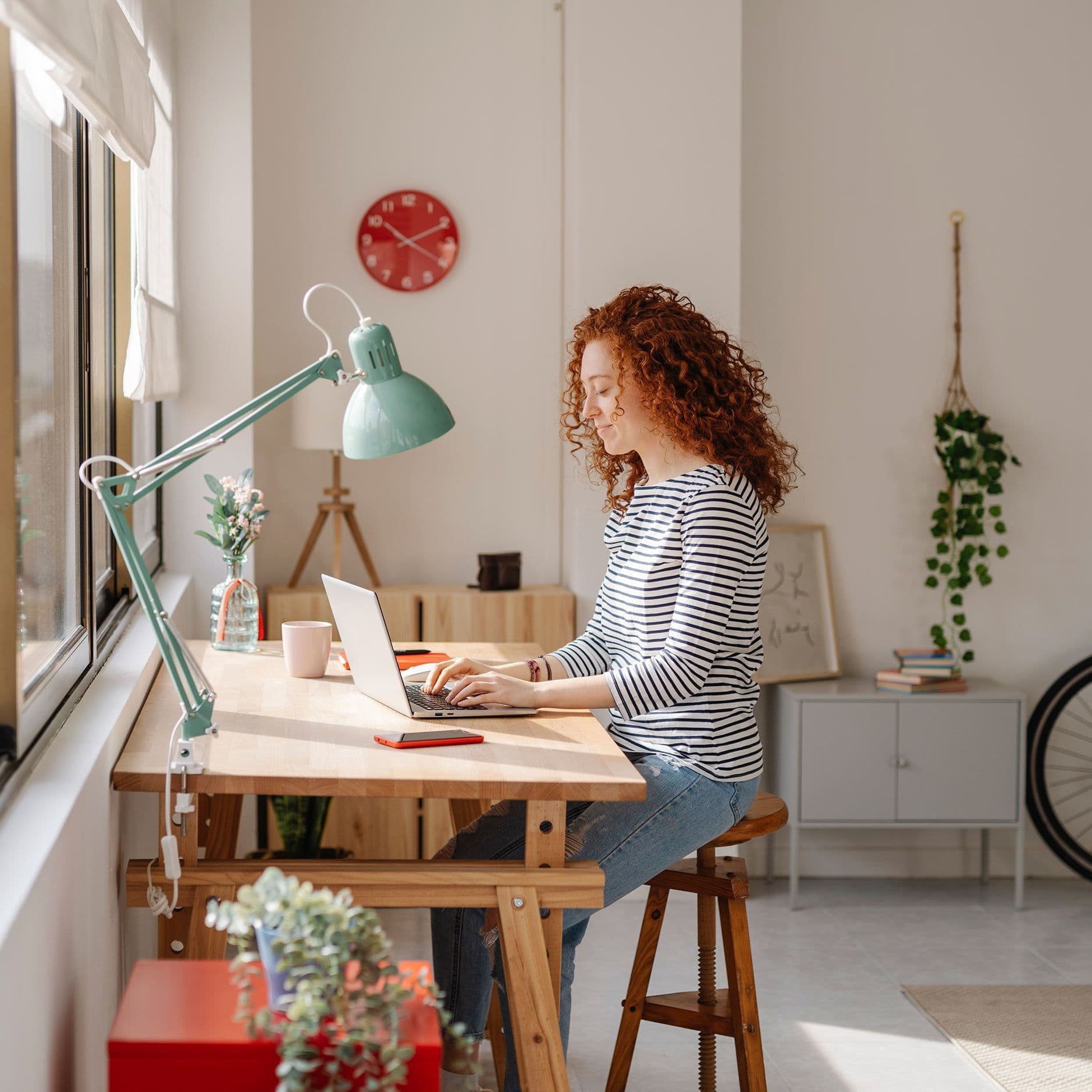 Eine junge Frau mit roten Locken arbeitet konzentriert an ihrem Schreibtisch in einem hellen, gemütlichen Homeoffice. Das Zimmer ist stilvoll eingerichtet mit modernen Möbeln, Pflanzen und dekorativen Elementen.
