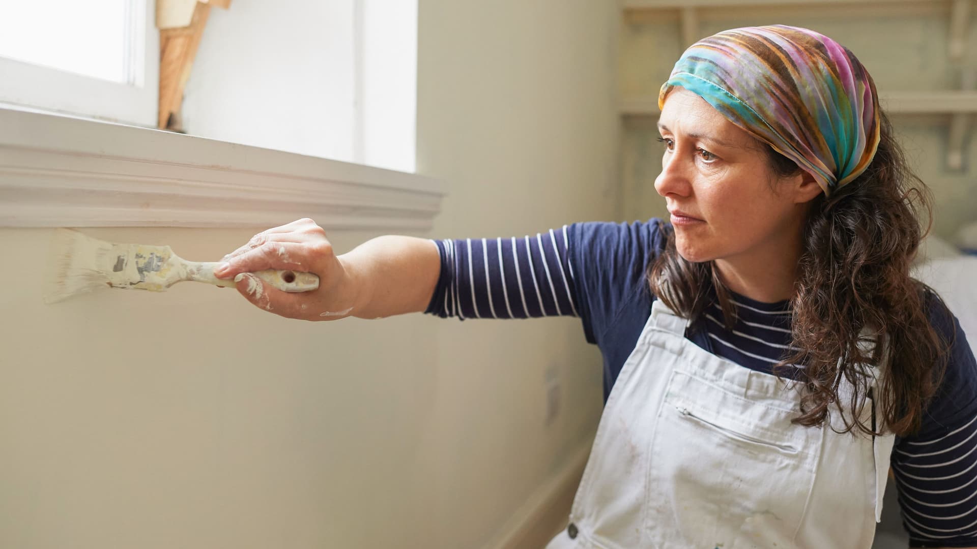 Frau in Latzhose streicht sitzend mit einem Pinsel eine weiße Wand unterhalb einer Fensterbank.