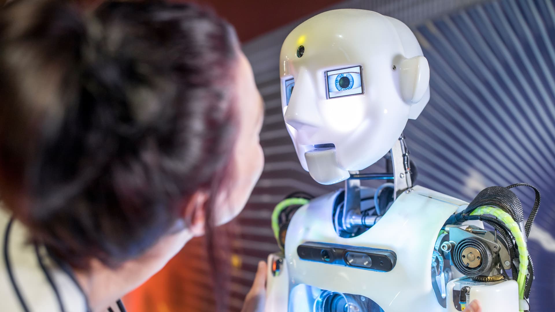 Technische Entwicklerin sieht sich humanoiden Roboter in der Werkstatt an. Er ist weiß und hat neben blauen Augen weitere blau leuchtende Elemente.