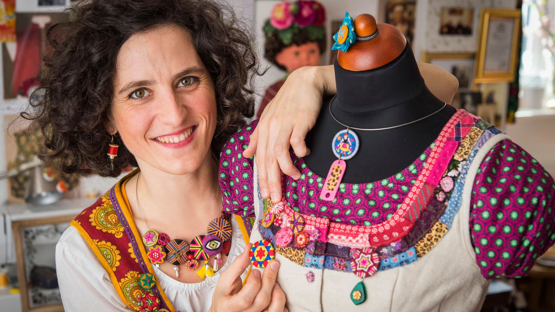 Junge Frau lächelt in die Kamera und hält einen Knopf an eine Schaufensterpuppe mit buntem Dirndl mit Posamentenknopf Collier in einem Atelier.