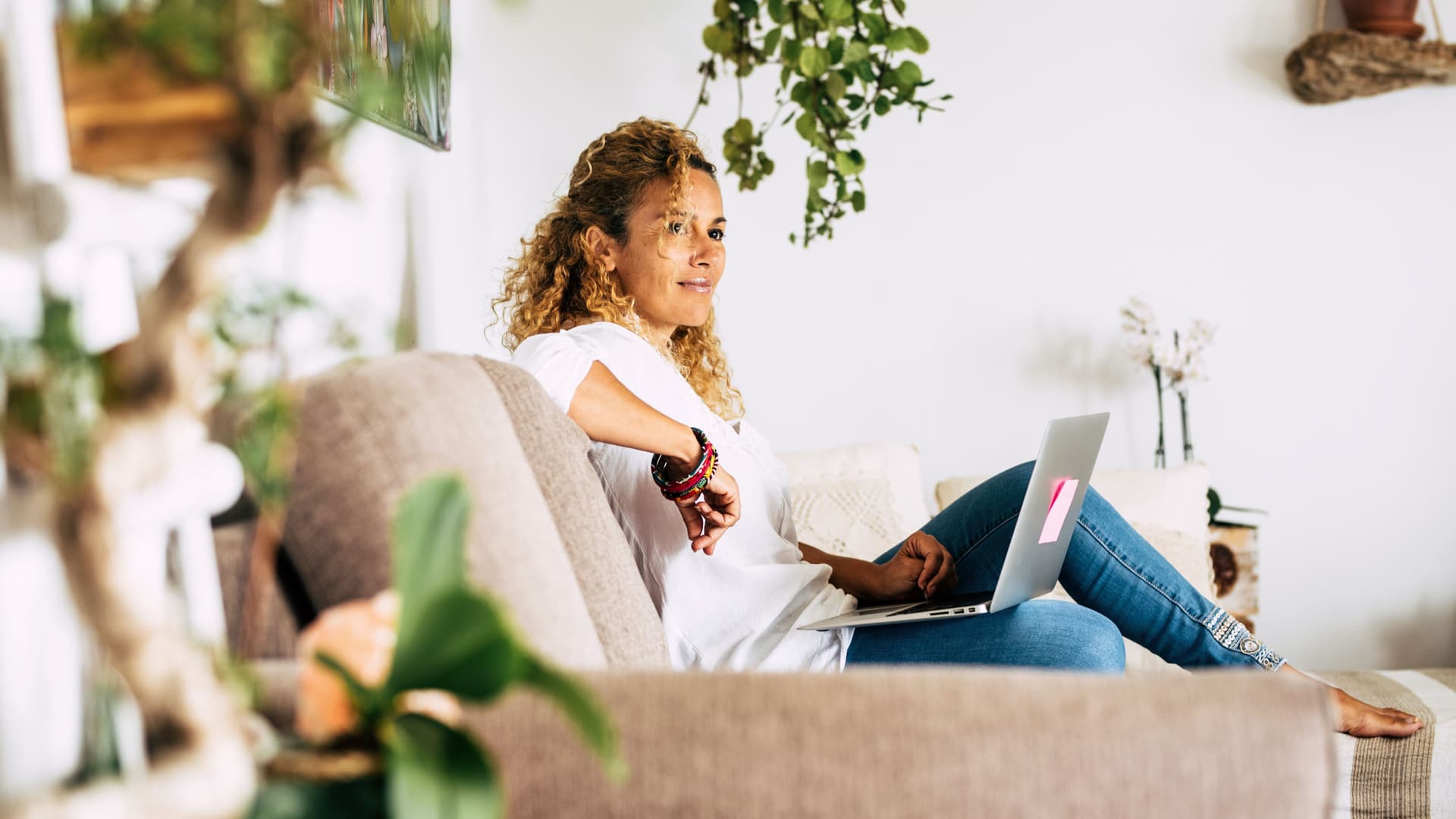 Junge Frau mit Locken sitzt entspannt auf einem Sofa umgeben von einigen Zimmerpflanzen. Sie hat einen Laptop auf ihrem Schoß.