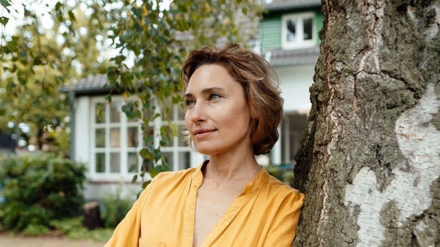Eine Frau lehnt an einem Baum im Garten vor ihrem Haus.