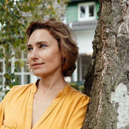 Eine Frau lehnt an einem Baum im Garten vor ihrem Haus.