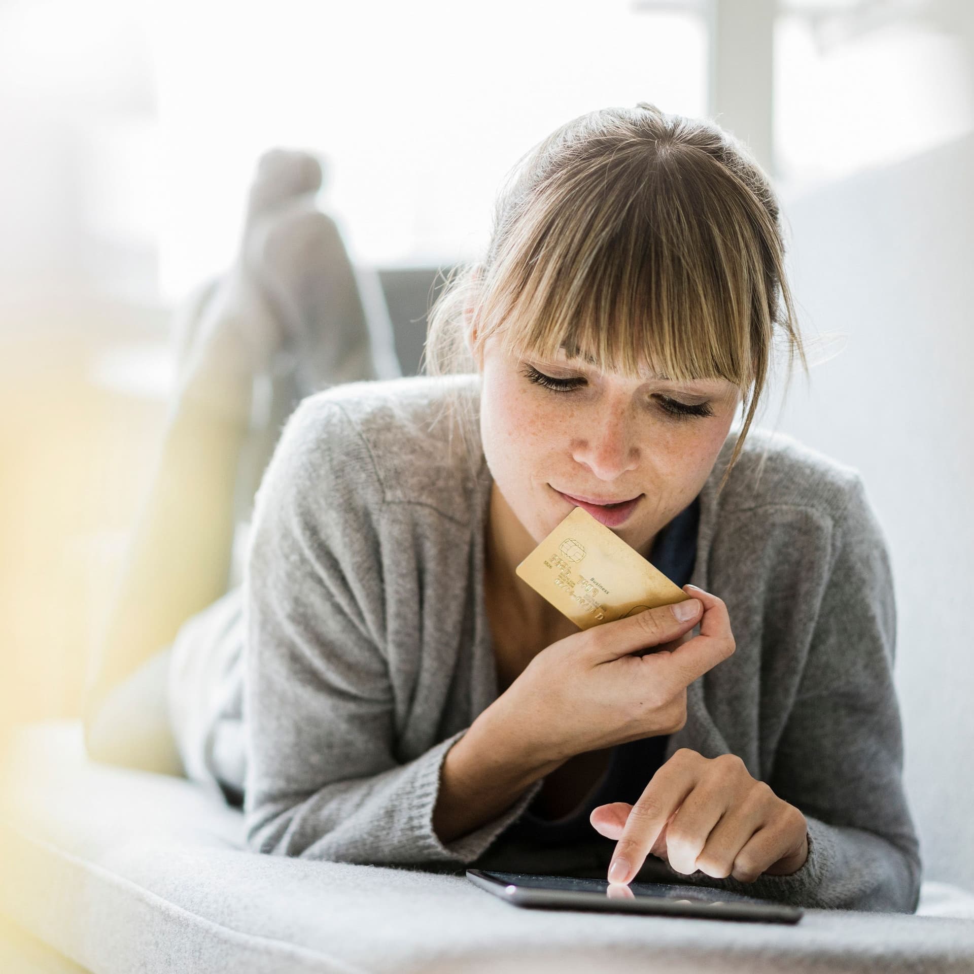 Junge Frau liegt mit ihrem Tablet auf einem Sofa, dabei hält sie eine Kreditkarte in ihrer rechten Hand.