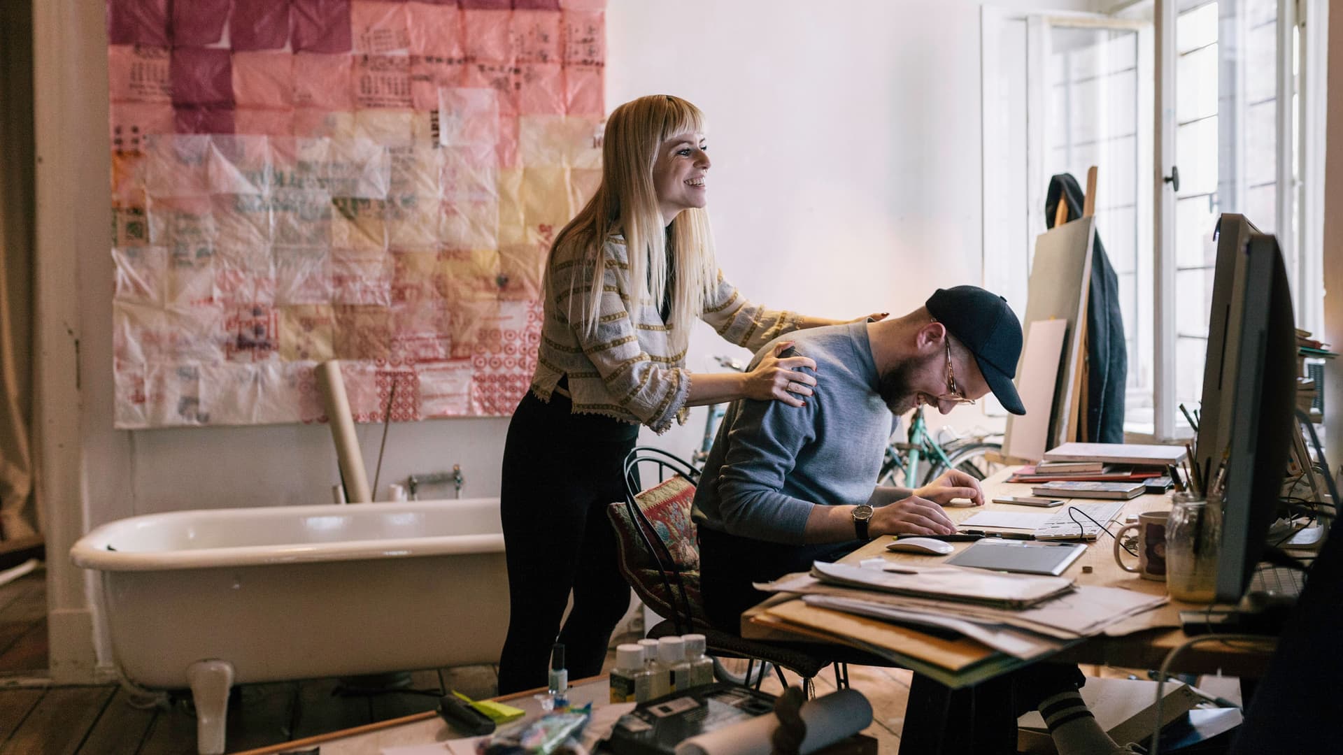 Ein Mann arbeitet im Home Office an einem Tisch. Eine Frau steht hinter ihm und hat die Hände auf seinen Schultern. Hinter den beiden steht eine Badewanne.
